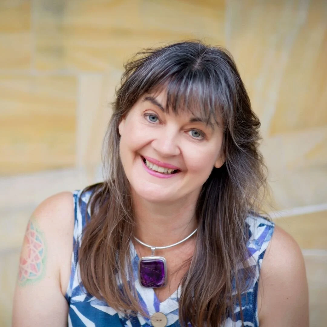 Portrait of a woman with shoulder-length brown hair, blue eyes, smiling, wearing a blue and white patterned sleeveless top, a purple pendant necklace, and a colorful tattoo on her right arm, with a wooden background.