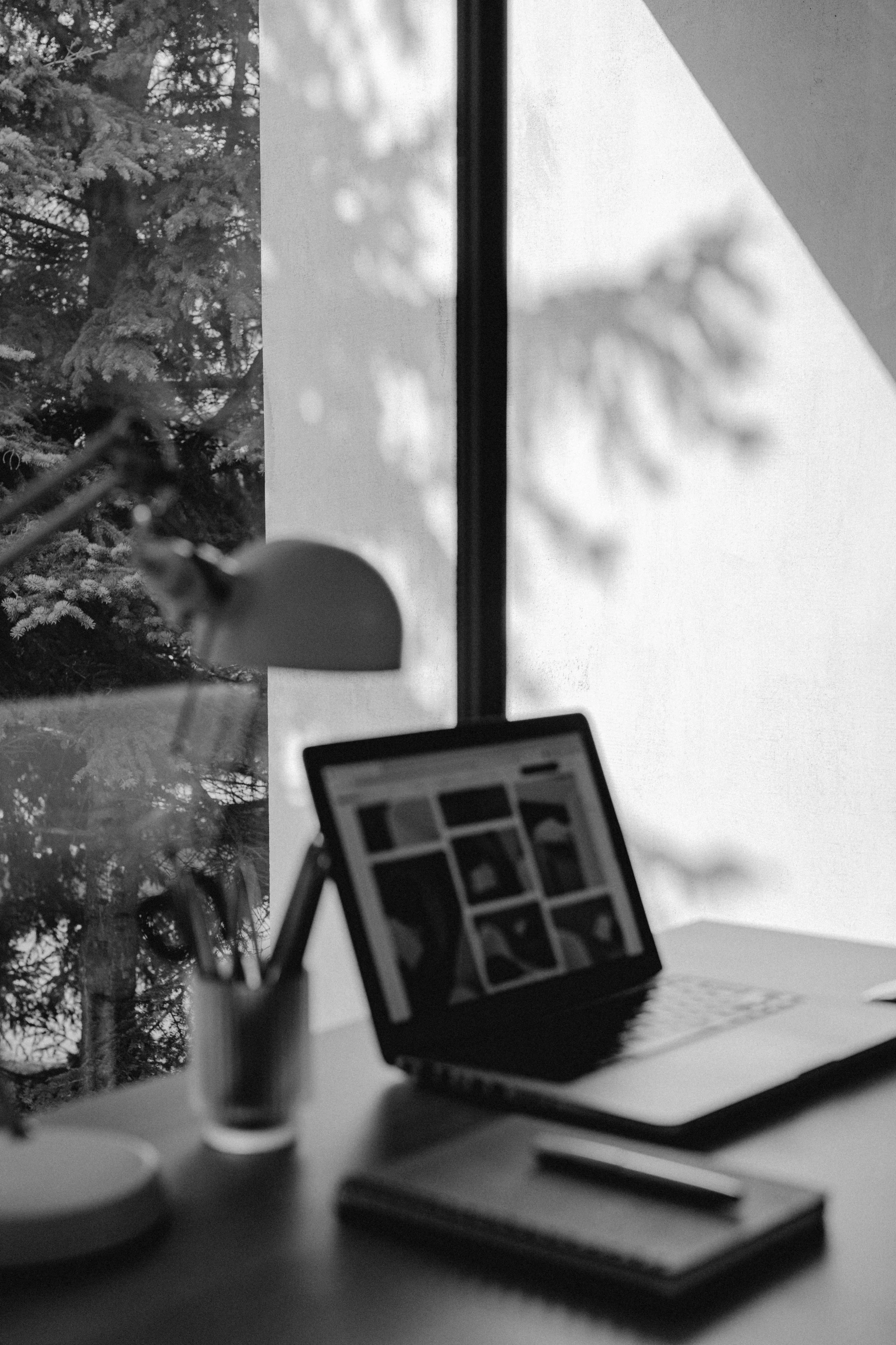 A black and white photo of a workspace with a laptop displaying a grid of images, a desk lamp, a notebook, and a pen. A large window shows outdoor trees and snow.