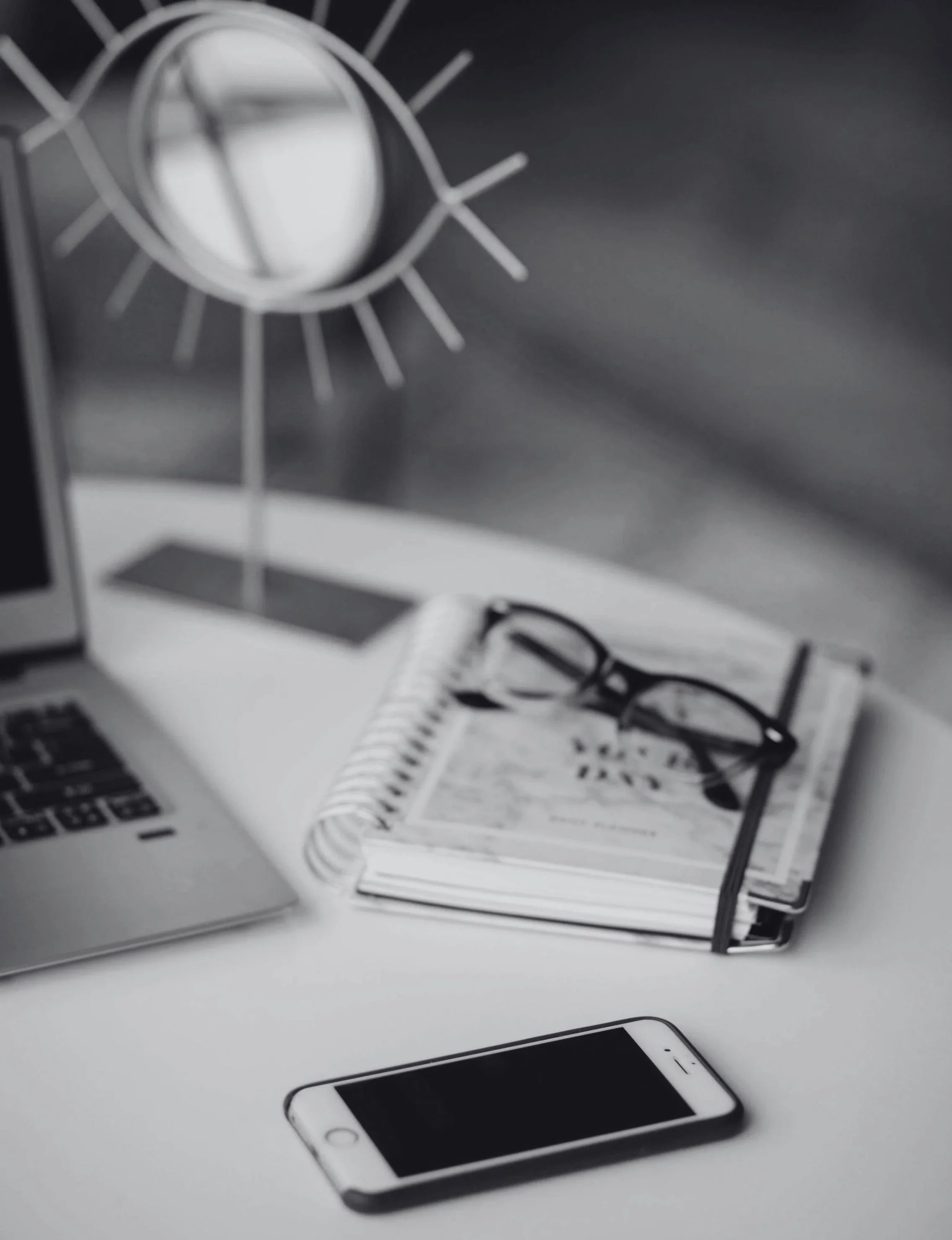 A workspace with a laptop, a smartphone, glasses, a spiral notebook, and a desk lamp.