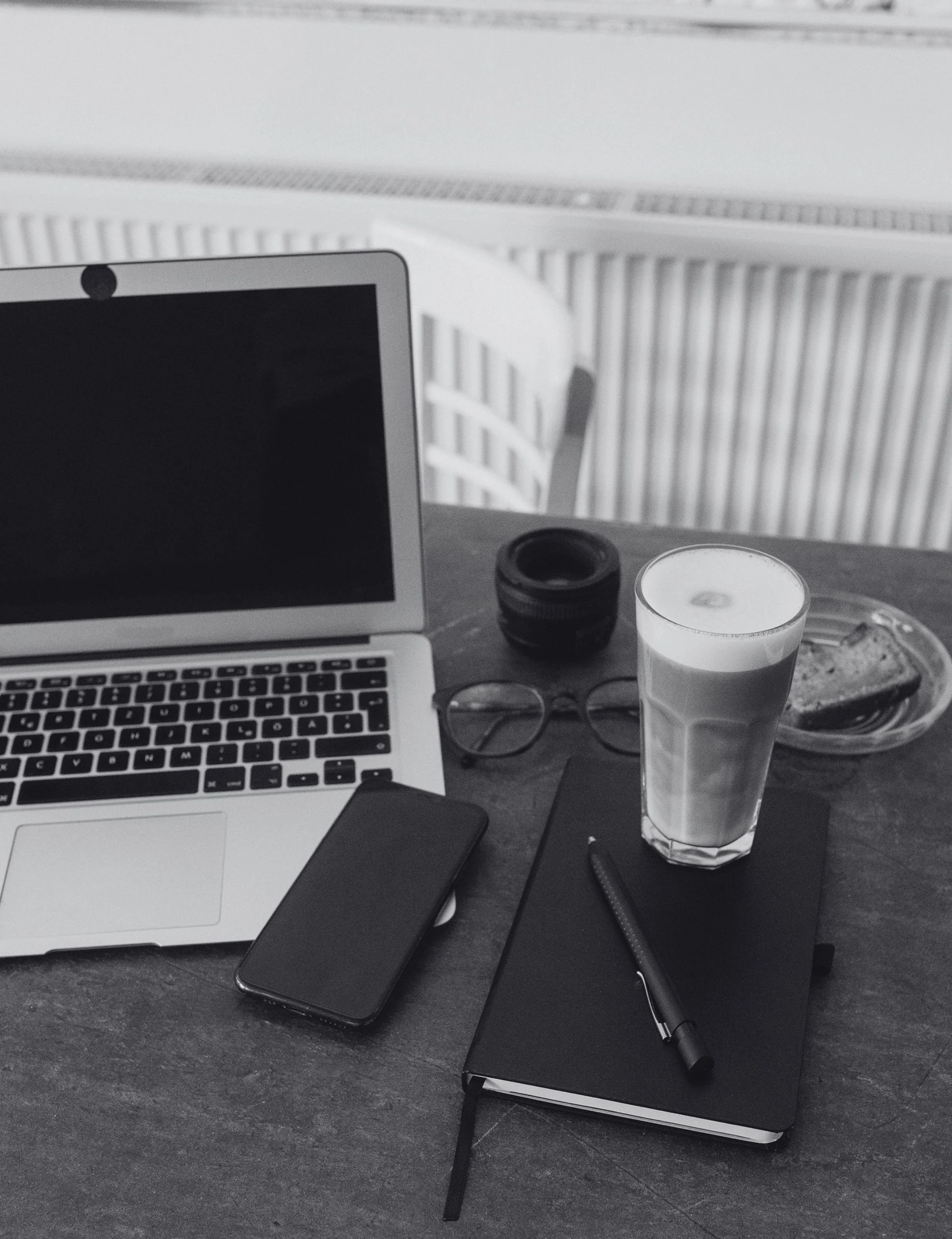 A work desk with a laptop, smartphone, glasses, notebook with pen, a glass of water, a small container, and a bowl of food, on a dark wooden surface with a white radiator and wall in the background.