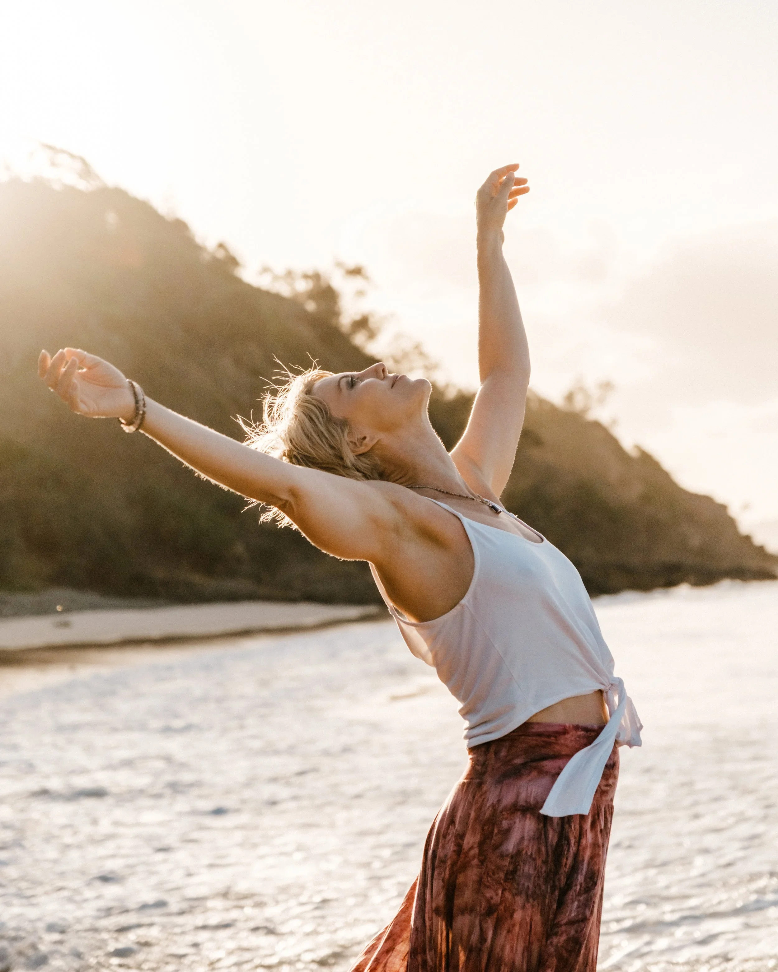 A woman with blonde hair dancing on a beach at sunset, with her arms raised and head tilted back.