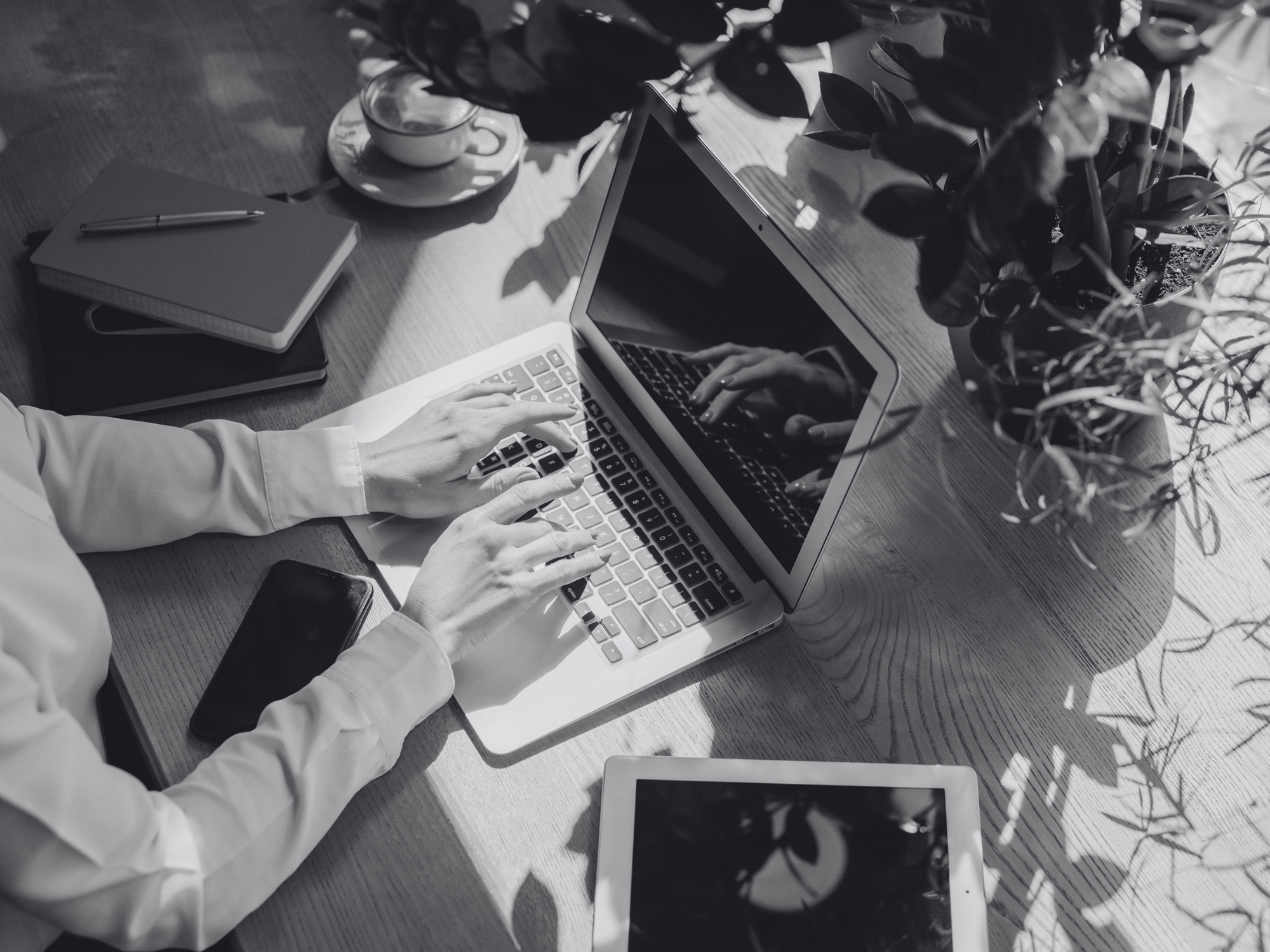 A person typing on a laptop at a desk surrounded by notebooks, a phone, a tablet, a cup, and a potted plant.