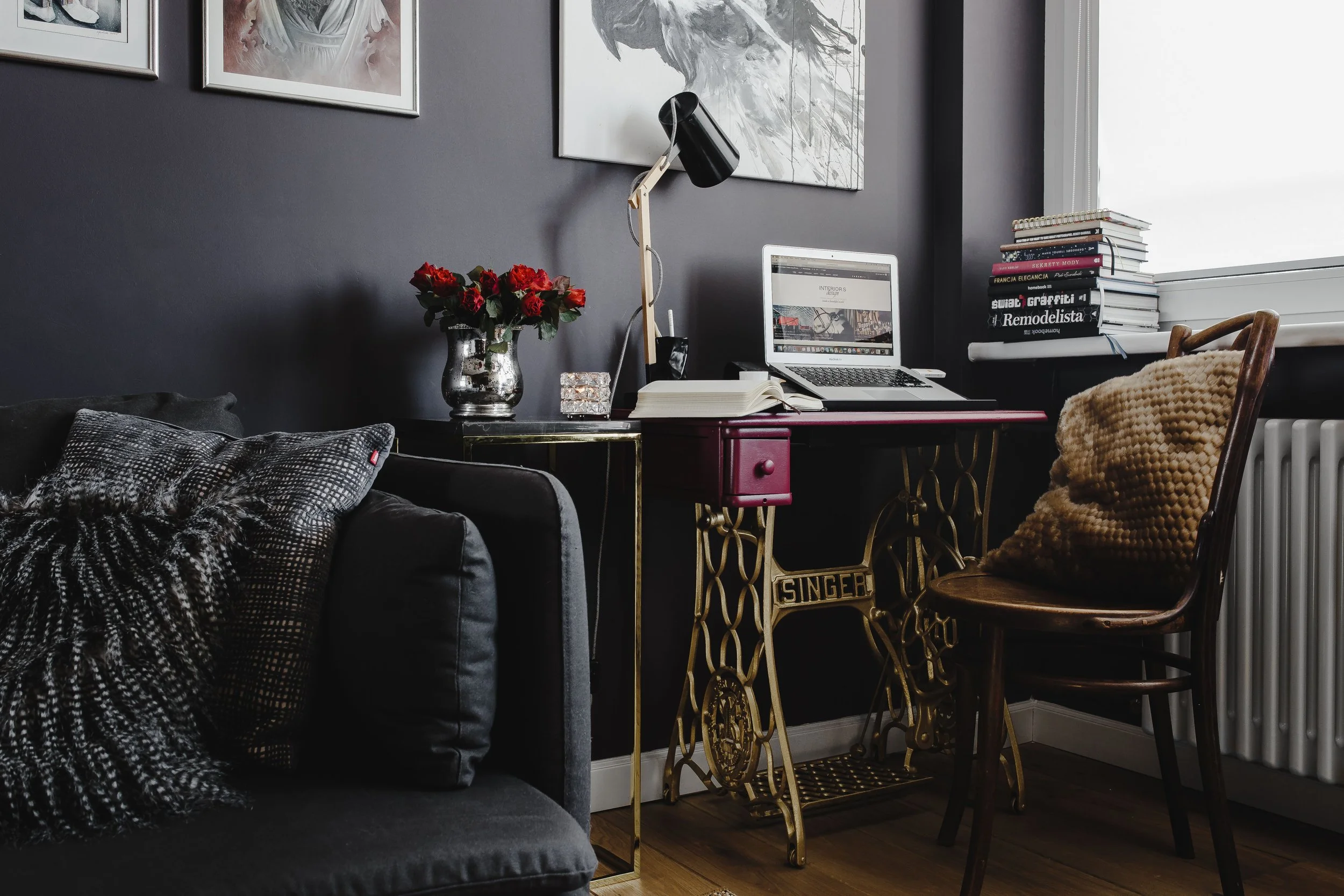 A home workspace with a vintage sewing machine table repurposed as a desk, a laptop, an open notebook, a black desk lamp, a vase of red roses, a glass of water, and a stack of books near a window. There is a dark wall with artwork and a cozy chair with a knitted pillow nearby.