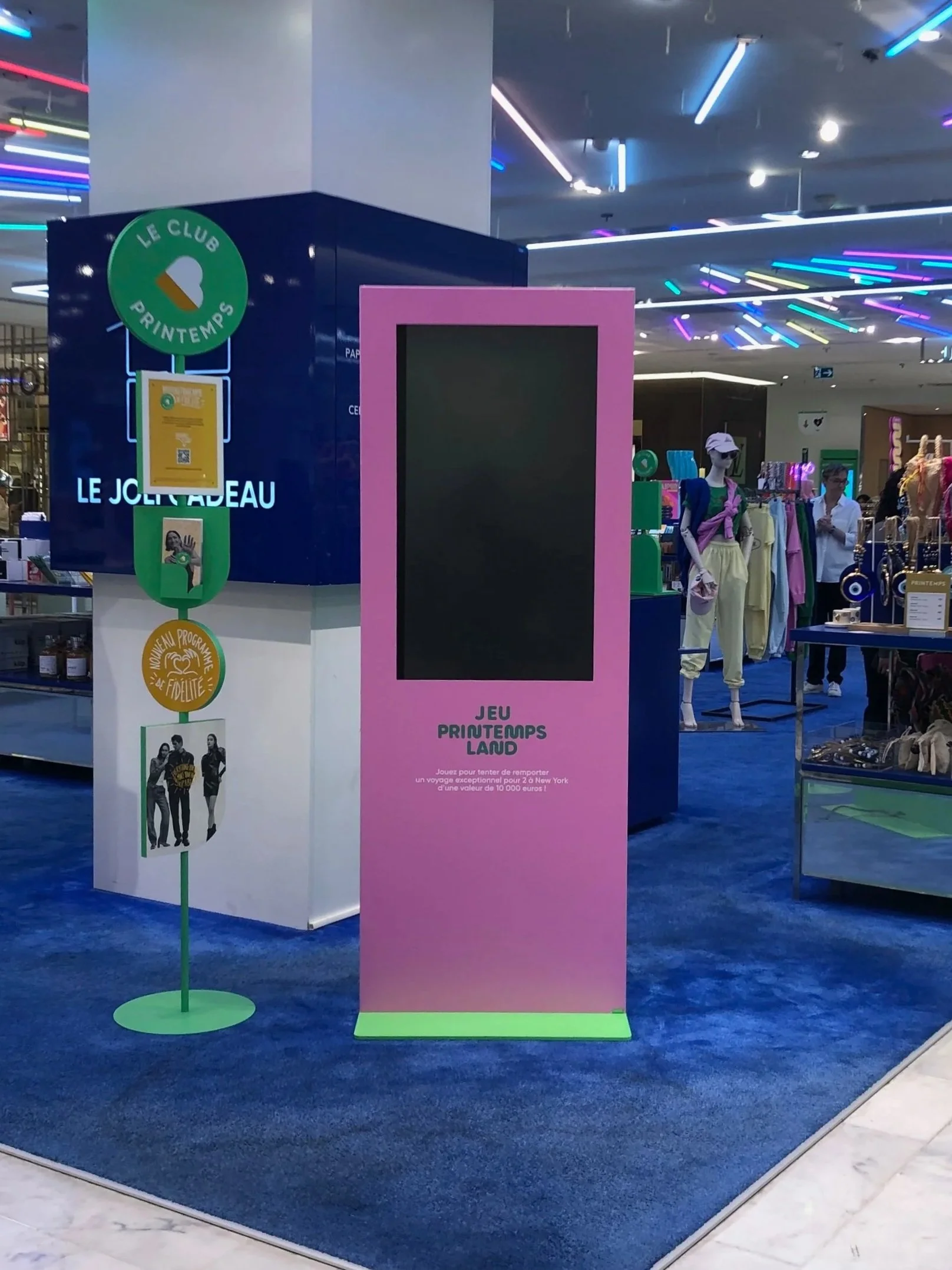 Pink digital kiosk on the store floor promoting the Printemps Land game, positioned near a "Le Club Printemps" loyalty program display and neon-lit ceiling installation.