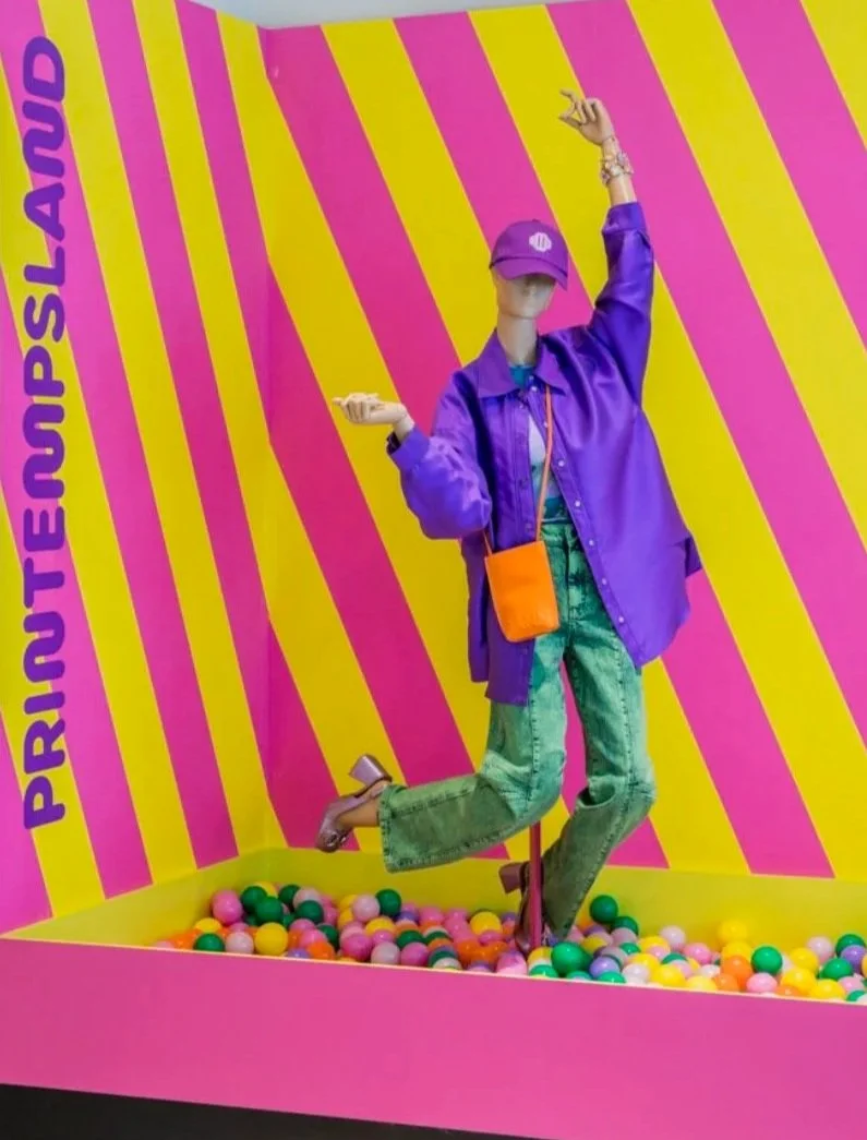Corner window display: a mannequin in purple and green outfit standing in a ball pit, against a vivid yellow and pink diagonal stripe backdrop with "Printempsland" in bold purple lettering.