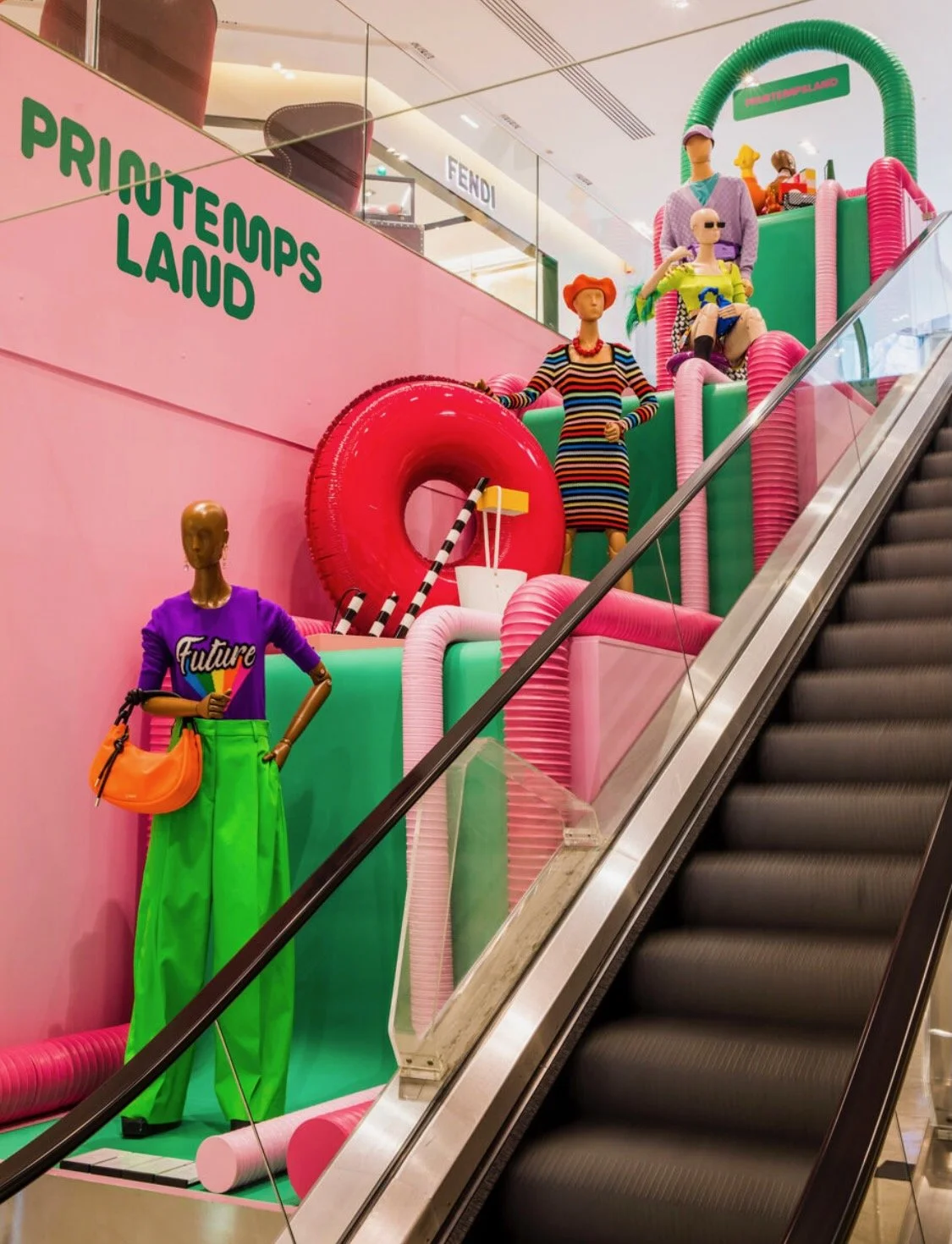 Escalator display featuring colorful mannequins styled in bold, neon outfits surrounded by inflatable pink tubes and props, with "Printemps Land" branding on a pink wall.