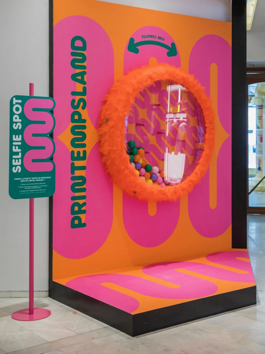 In-store selfie activation spot featuring an orange feather-framed circular mirror/lottery drum on a graphic pink-and-orange wall, with a "Selfie Spot" sign inviting customers to participate and win gift vouchers.