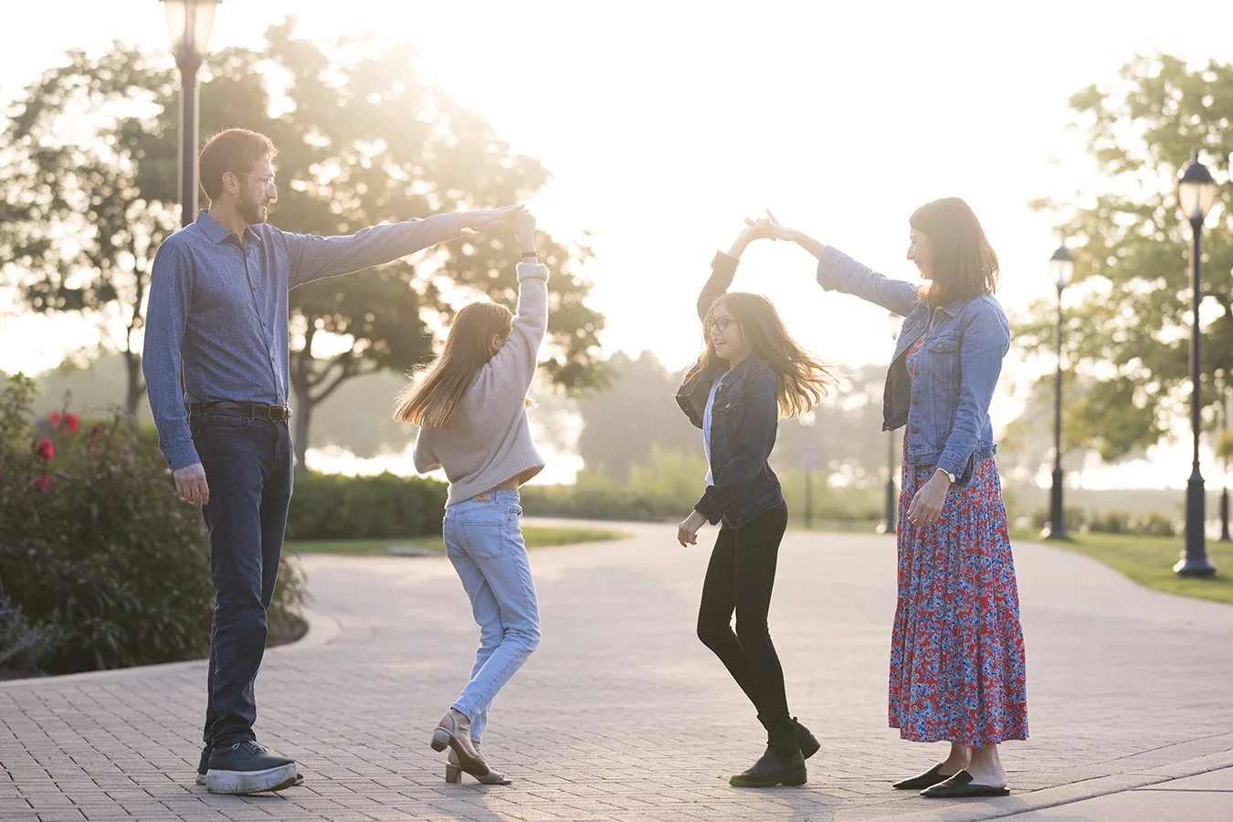 Family dancing outdoors on a sunlit pathway, with parents holding hands above two children during a playful twirl, surrounded by trees and lampposts.