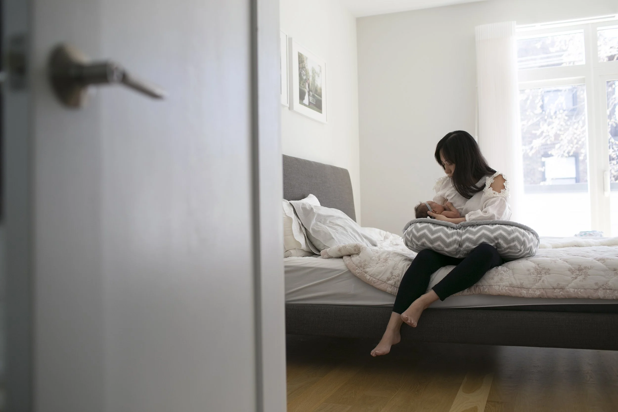 Woman sitting on a bed breastfeeding a baby in a bright room.