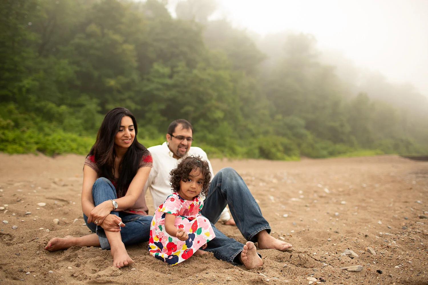 A family sitting on a sandy beach with greenery in the background on a foggy day. The child is wearing a colorful dress and holding sand.