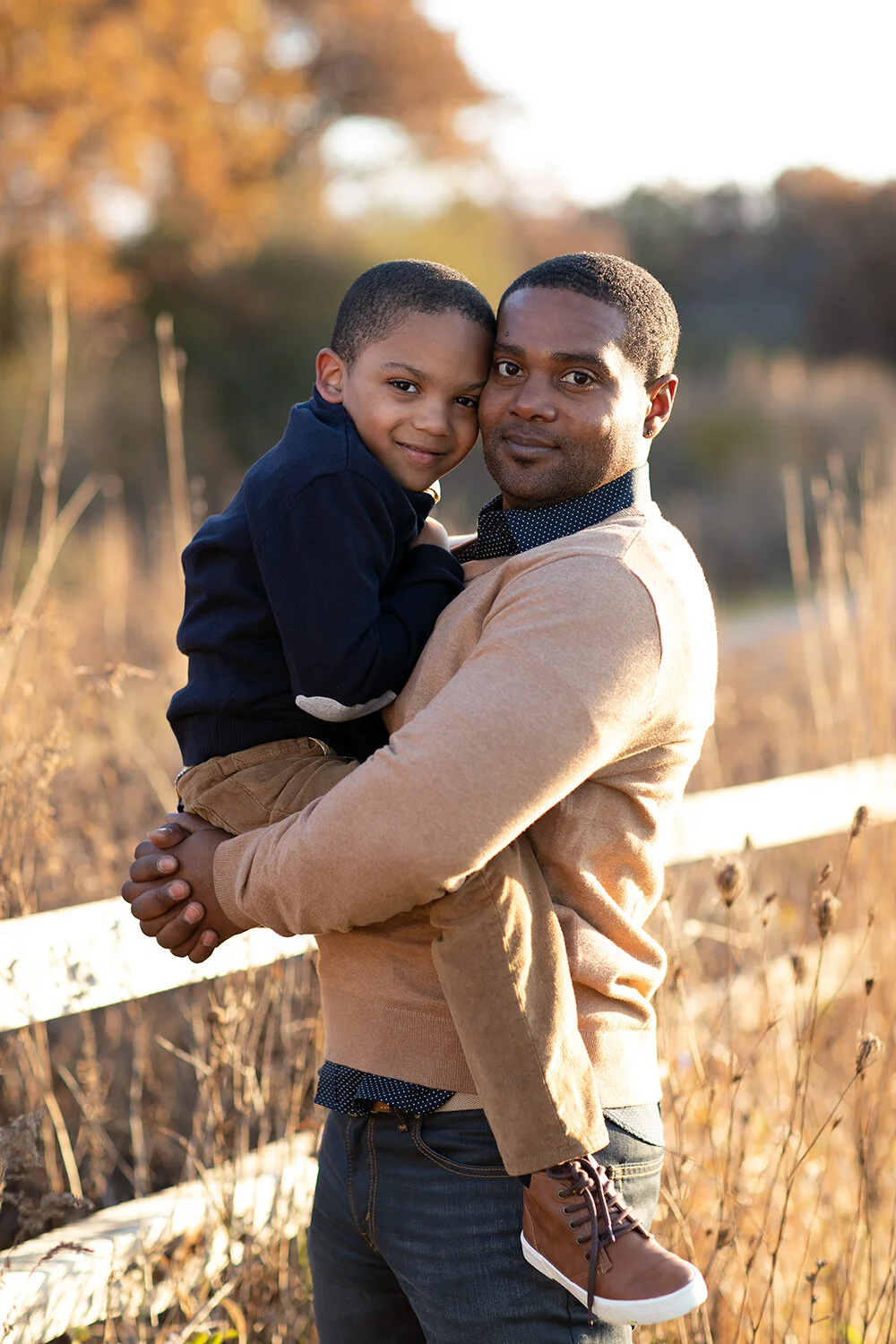 A man holding a young boy in an outdoor setting with autumn foliage.