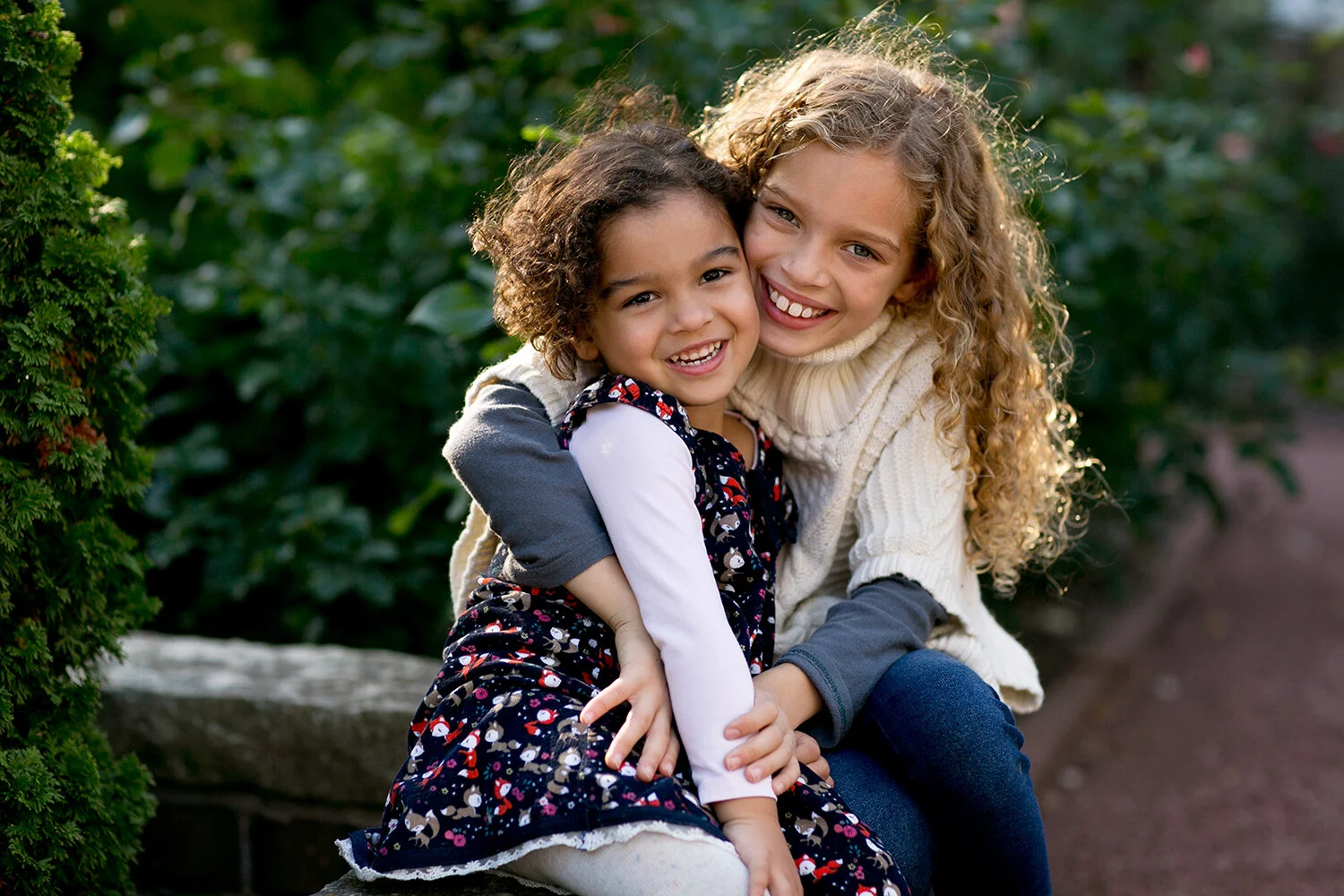 Two smiling young girls sitting together in a park, one with curly blonde hair wearing a sweater, and the other with short brown hair in a floral dress, surrounded by greenery.