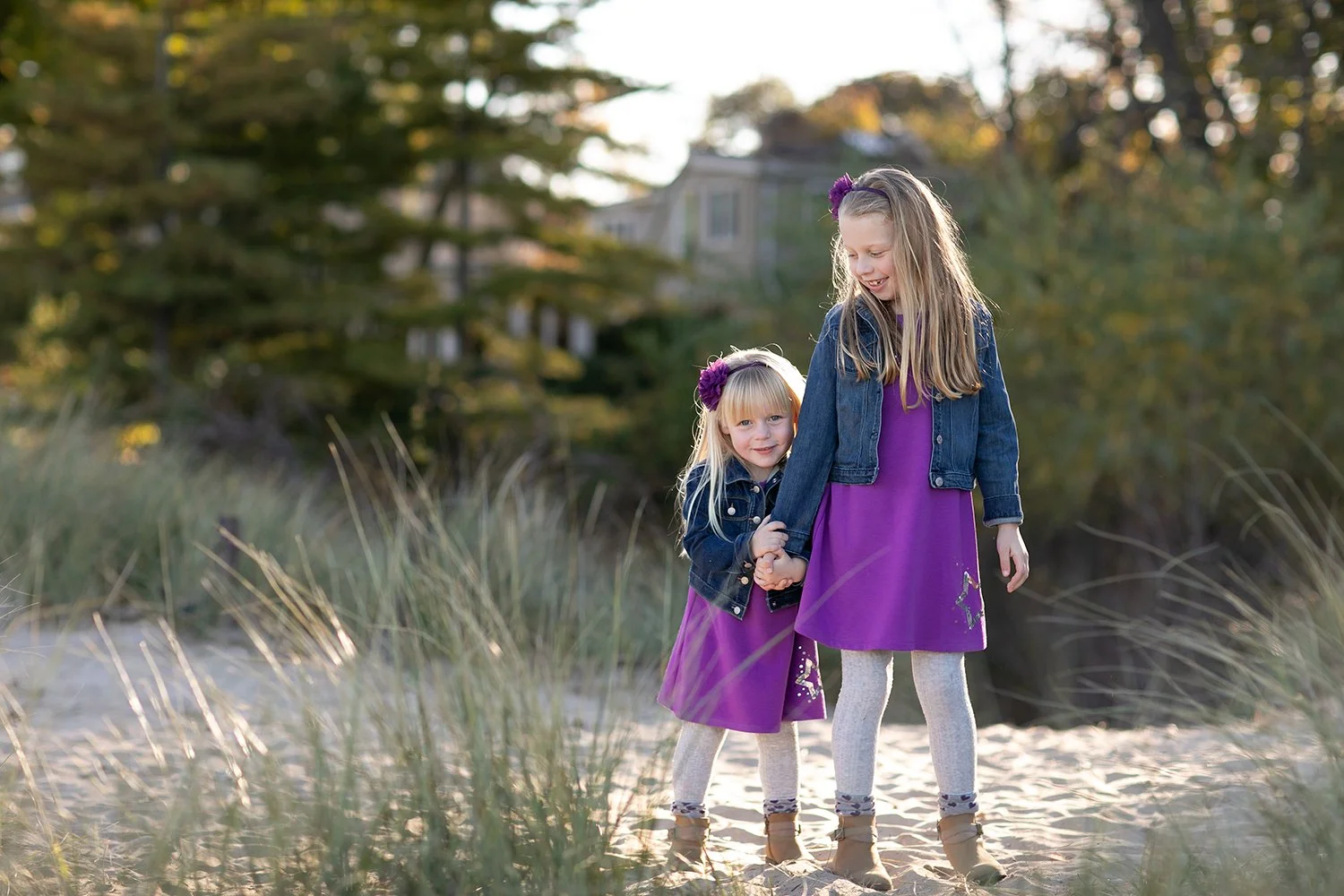 Two young girls in purple dresses and denim jackets stand together on a sandy path with grass and trees in the background.