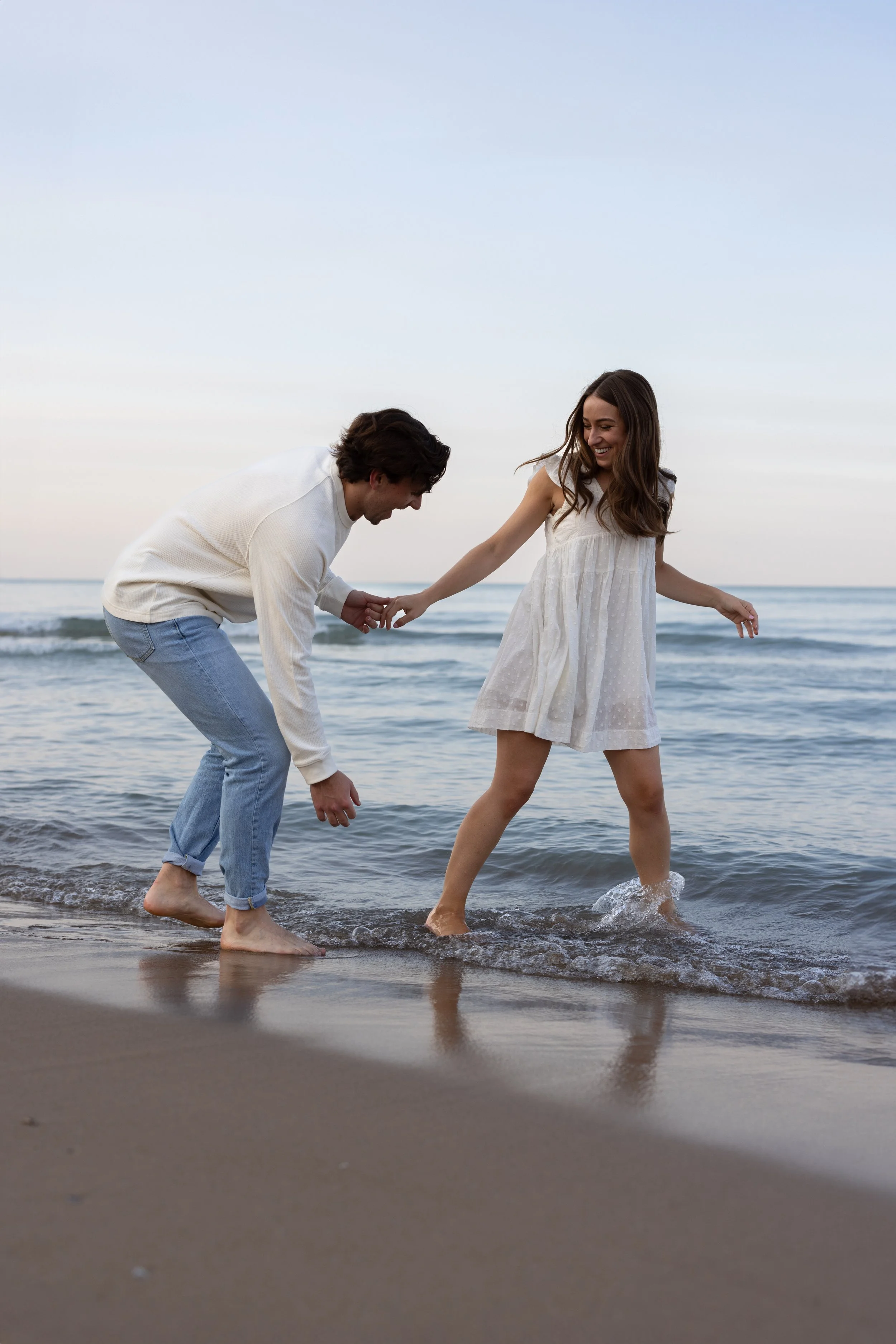 A smiling couple playing on a sandy beach with ocean waves in the background, wearing casual white and light blue clothing.