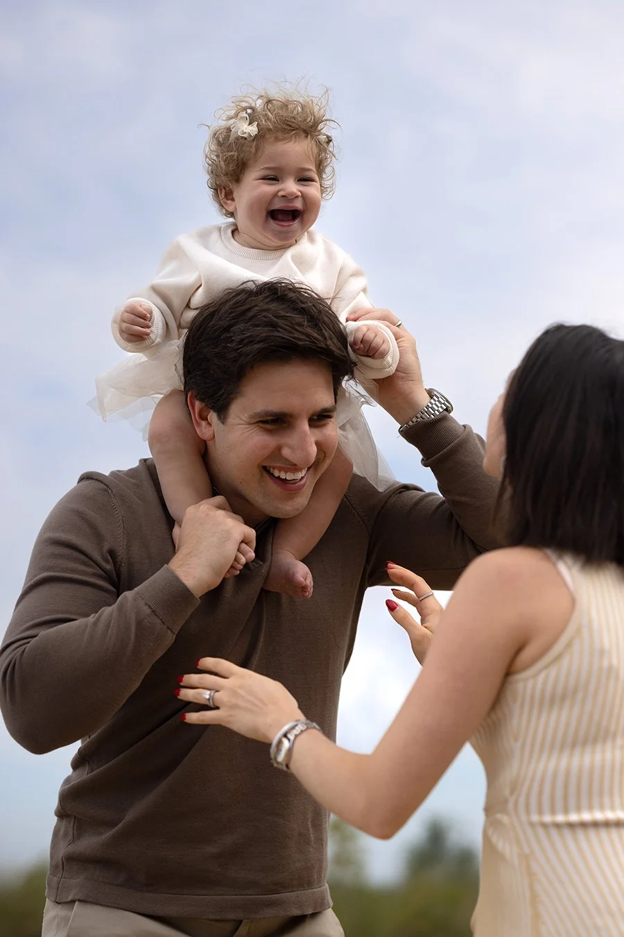 Happy child sitting on a man's shoulders with a woman nearby, outdoors.
