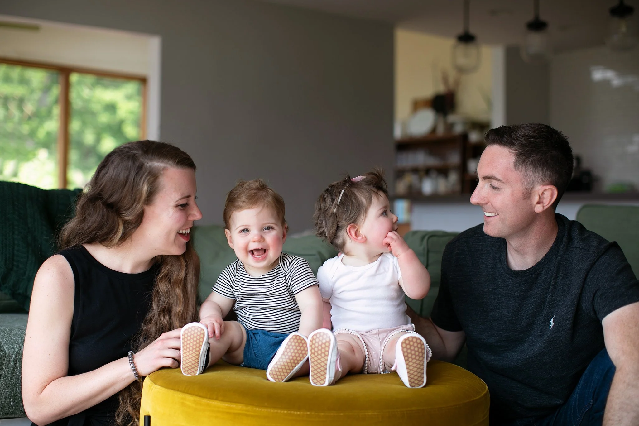 A cheerful family with two toddlers sitting indoors on a yellow ottoman, surrounded by a man and a woman.