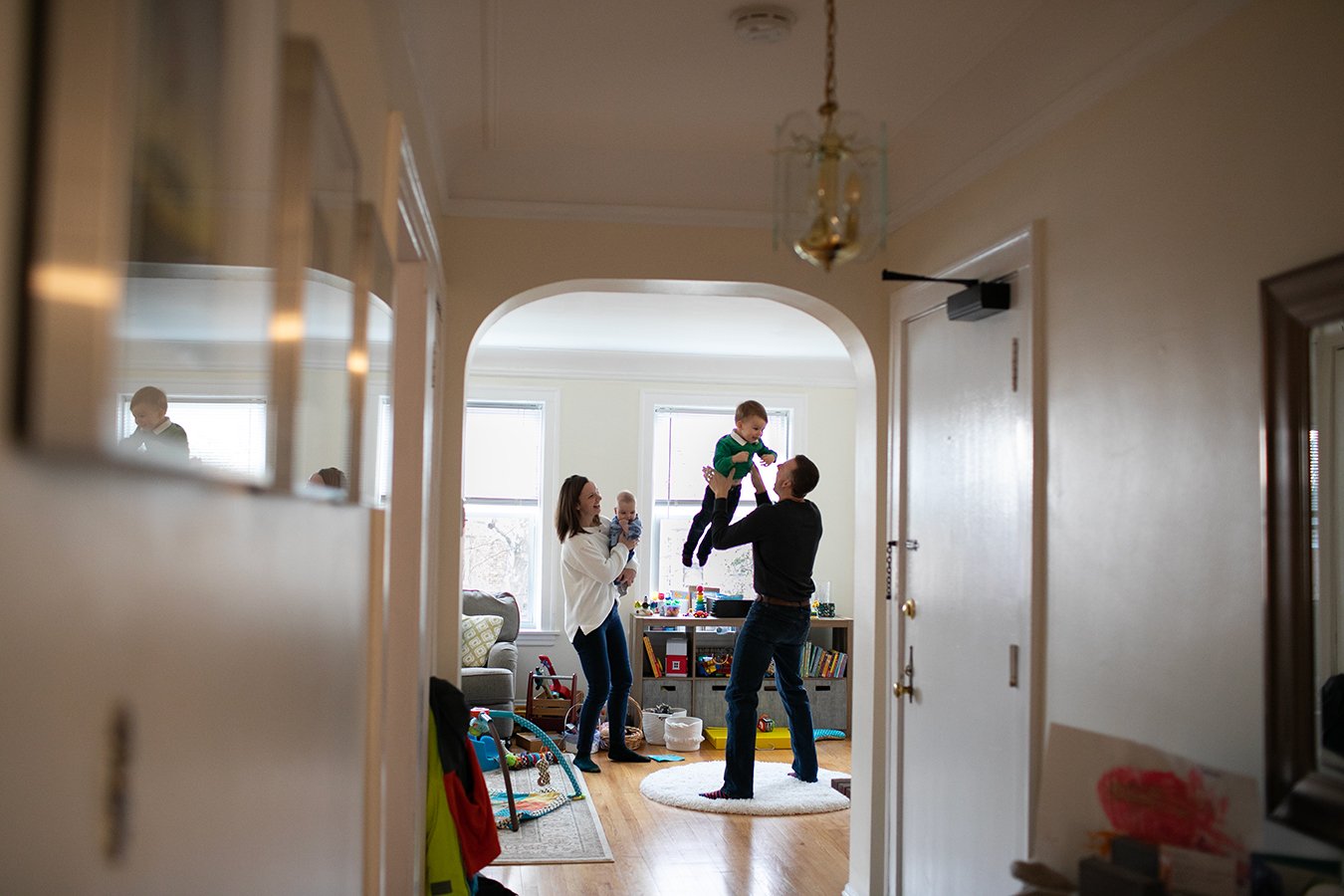 Family in living room; father lifting child; mother holding baby; toys on floor.
