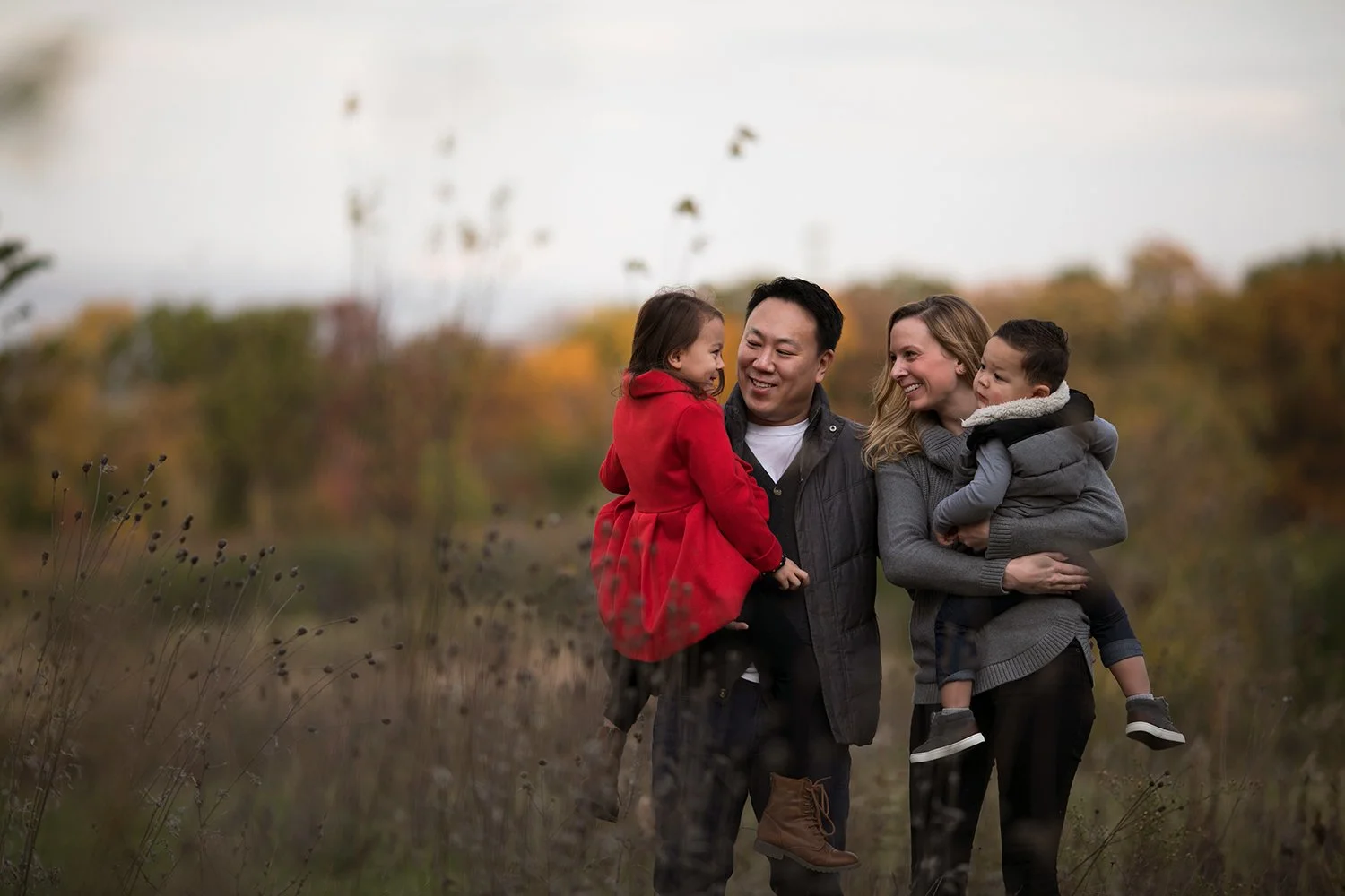 A family of four in an autumn field, with parents holding young children, smiling and wearing fall clothing.