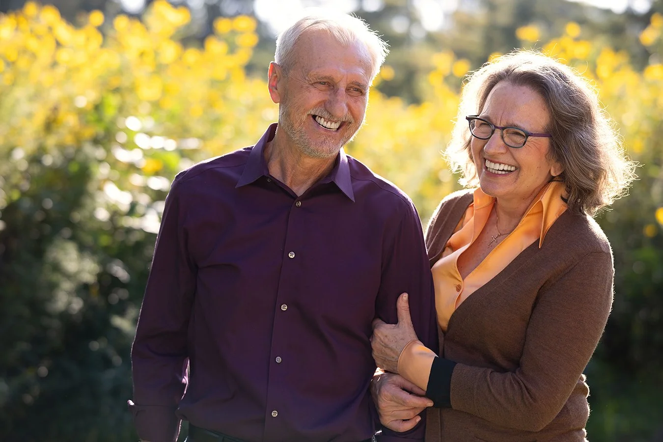 Smiling elderly couple embracing outdoors in a garden with yellow flowers.