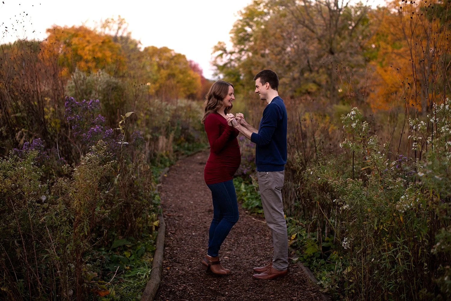 An expecting couple holding hands and facing each other on a path surrounded by autumn foliage.