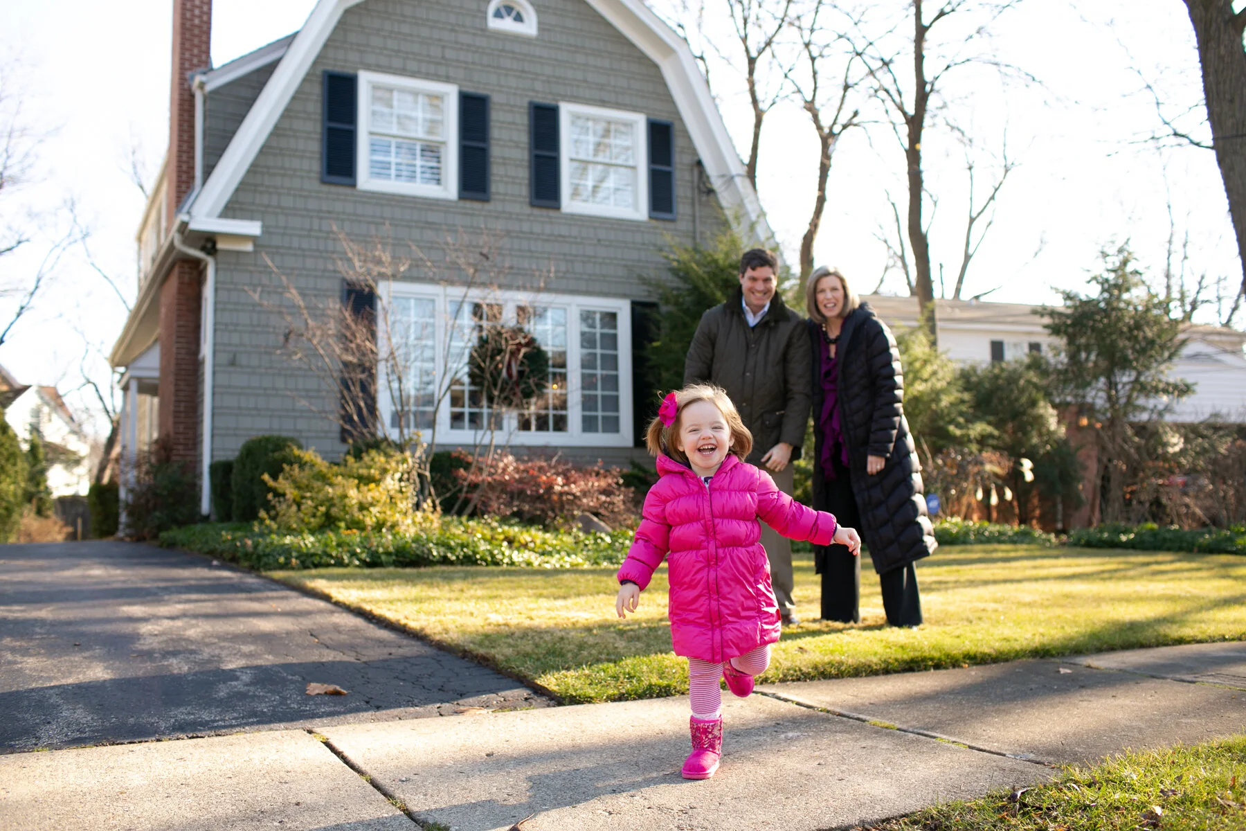 A young girl in a bright pink coat runs joyfully on a sidewalk with a smiling man and woman, presumably her parents, standing behind her. The family is in front of a suburban house with gray siding and black shutters, surrounded by winter foliage. The setting appears to be a sunny day.