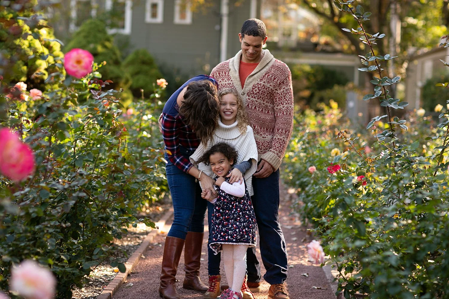 A family of four standing in a rose garden, smiling and embracing each other. The garden is filled with blooming pink and red roses. Trees and a building are in the background.