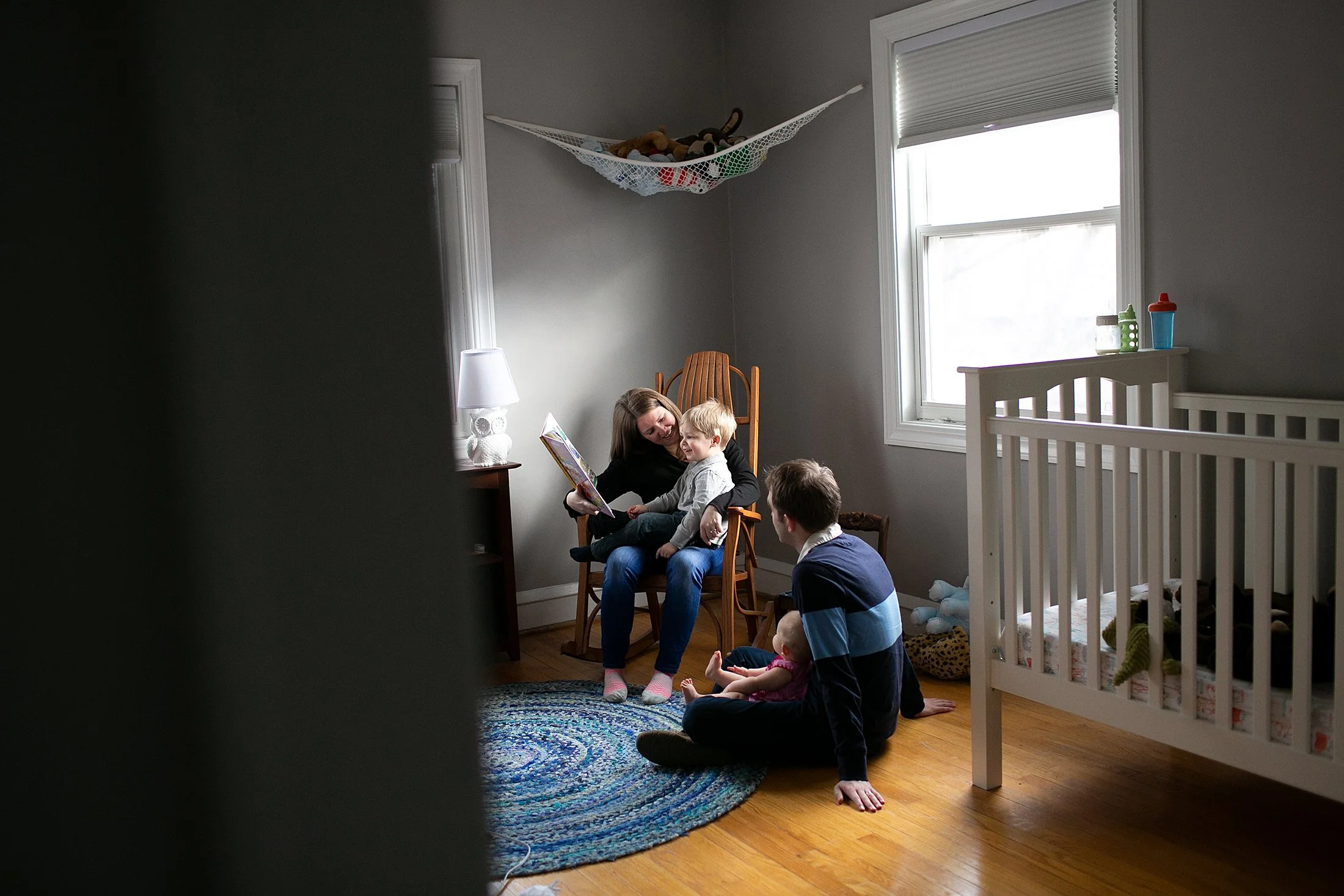 A family sitting in a nursery with a woman reading a book to a child on her lap, a man sitting on the floor with a baby, a crib, and a toy hammock with stuffed animals.