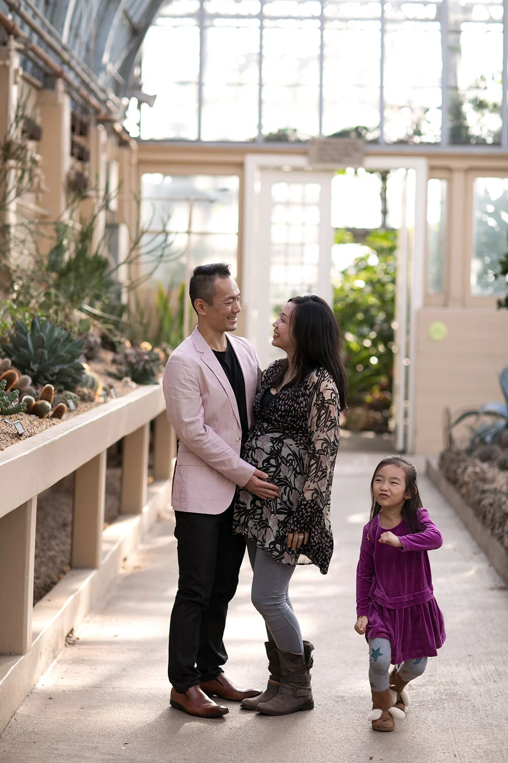 Family in a greenhouse with plants, man in pink jacket, woman in floral dress, child in purple dress.
