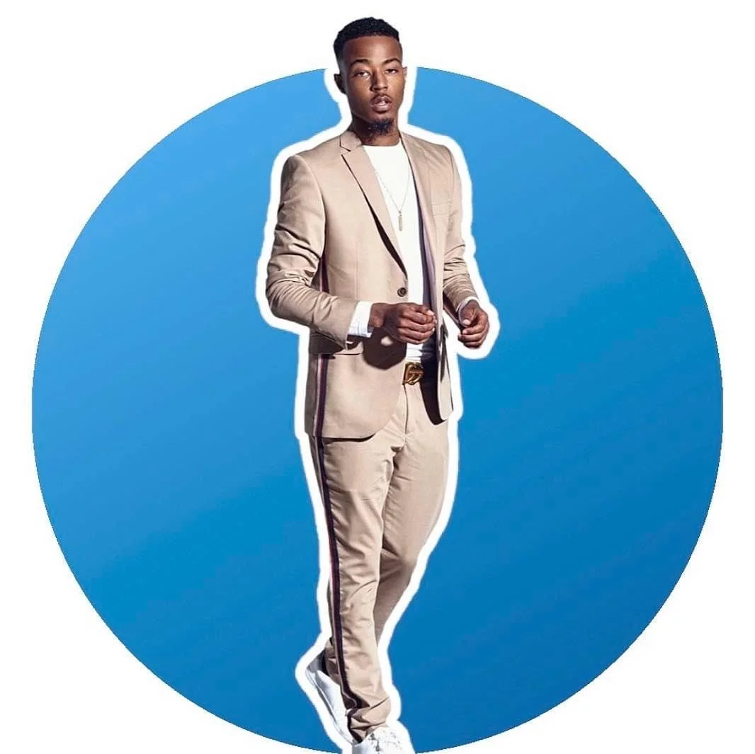 A young man in a beige suit with black and white trim, white sneakers, and a chain necklace standing against a blue circular background.