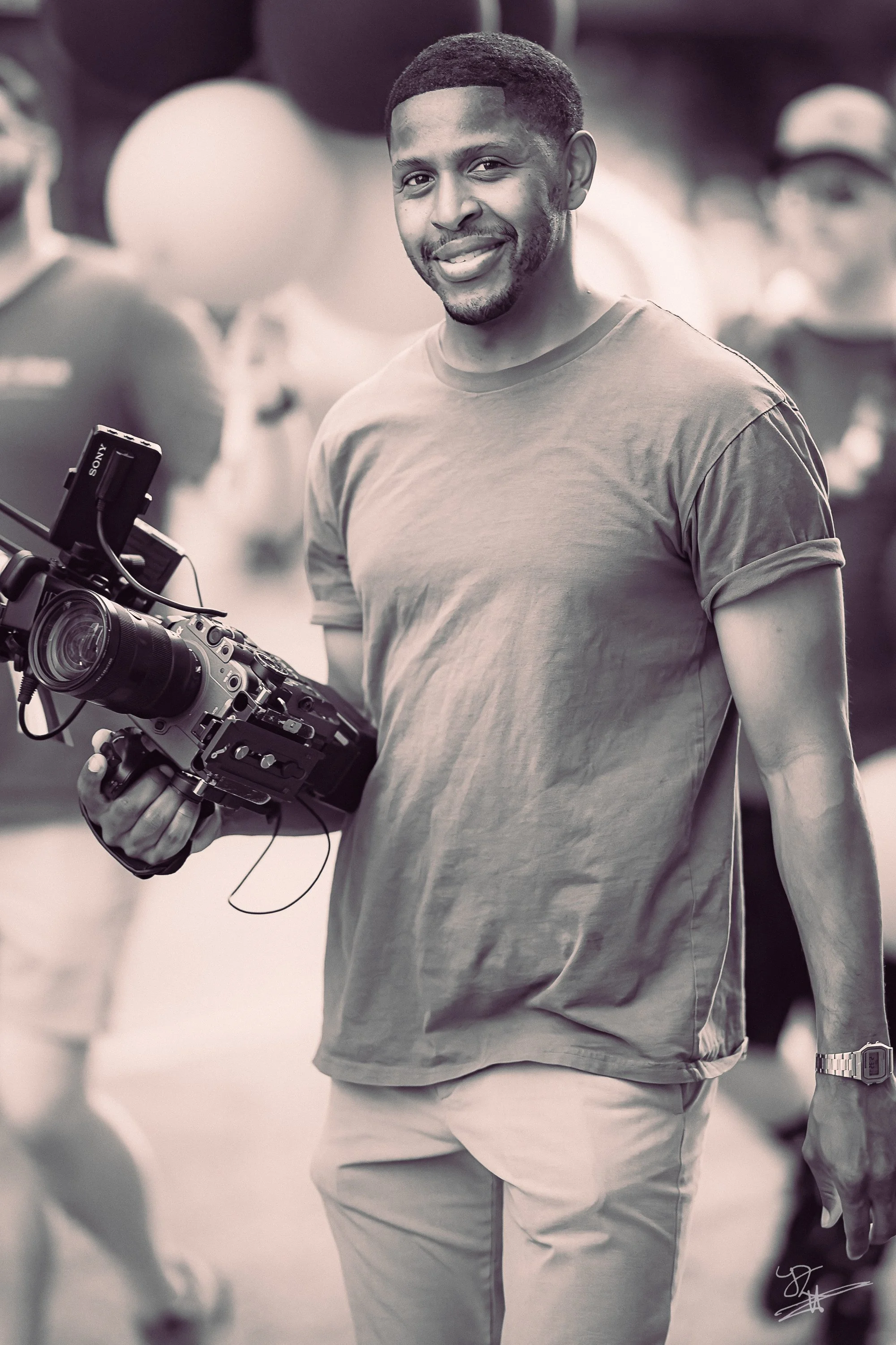 A man holding a professional video camera, smiling, standing in an outdoor event with balloons and people in the background.