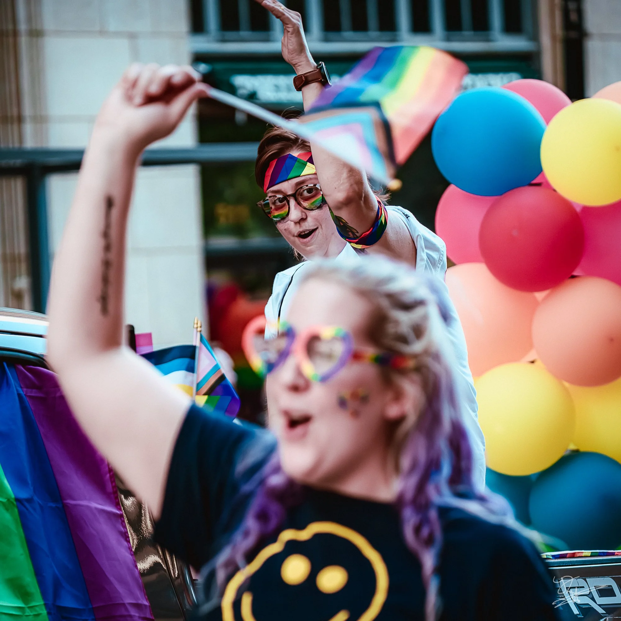 Two women celebrating at a pride parade, adorned with rainbow-colored accessories, sunglasses, and face paint, standing in front of a colorful balloon backdrop.