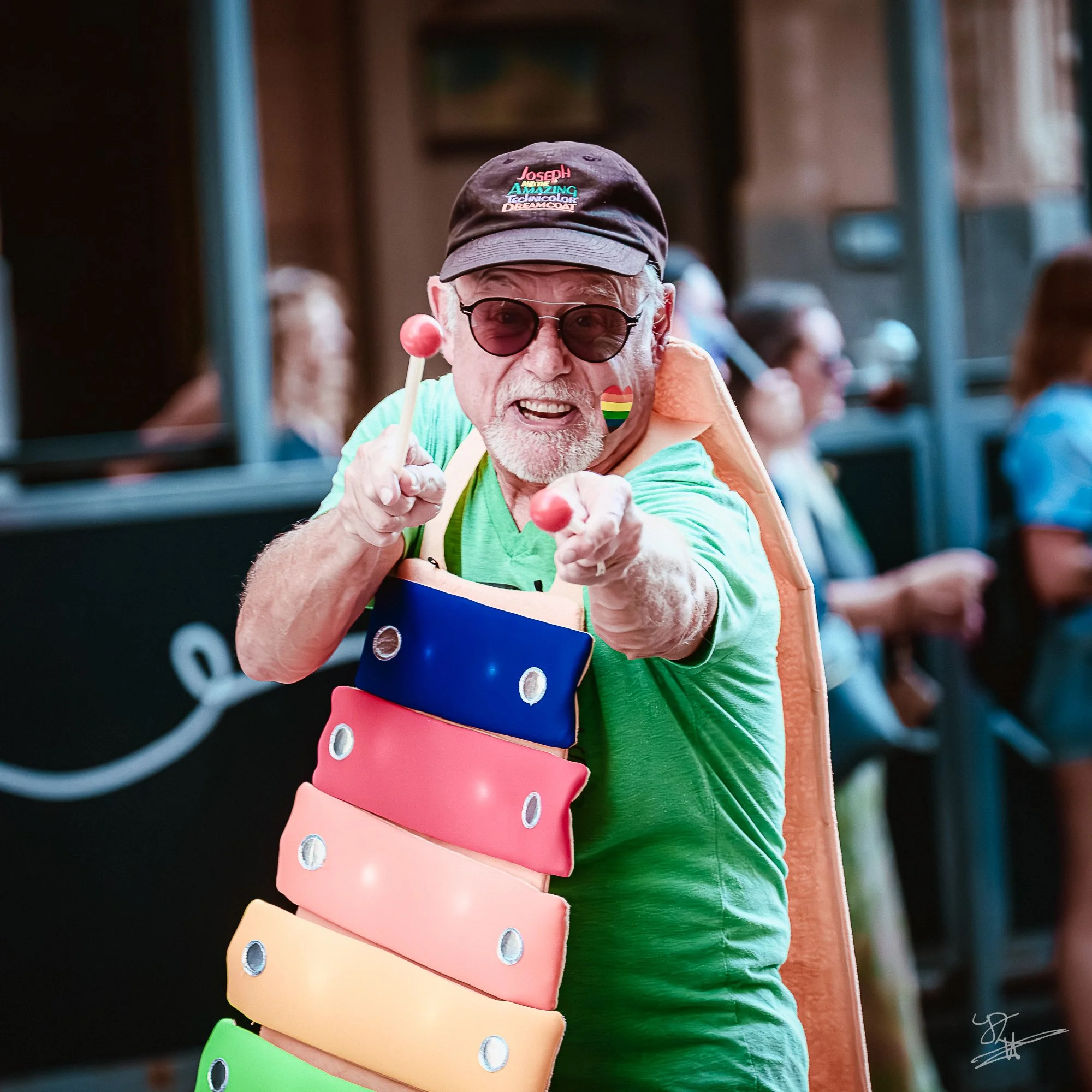 Older man in sunglasses and a cap playing a colorful xylophone at a lively event.