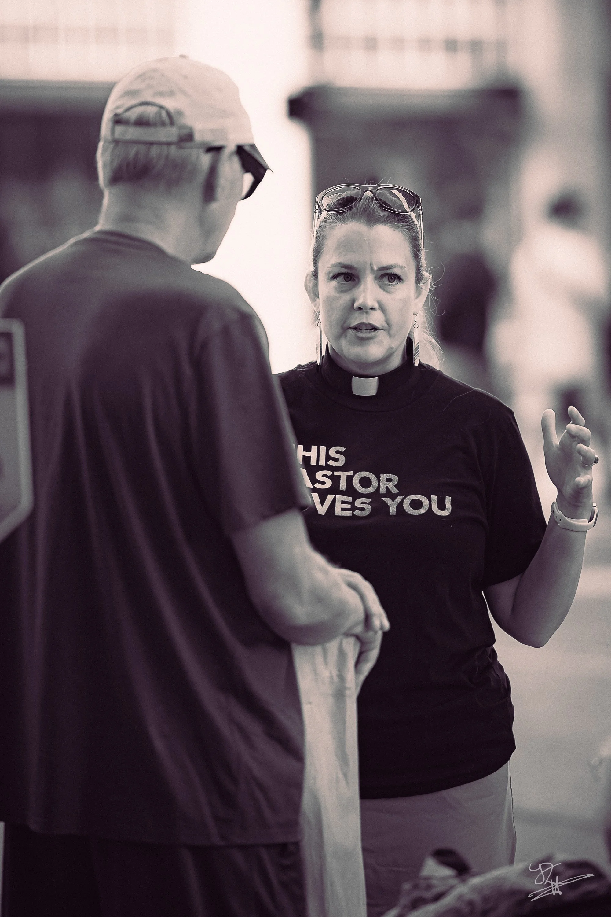 A woman with a clerical collar talking to a man in sunglasses and a cap, with a sign on her shirt that reads 'This pastor loves you'.