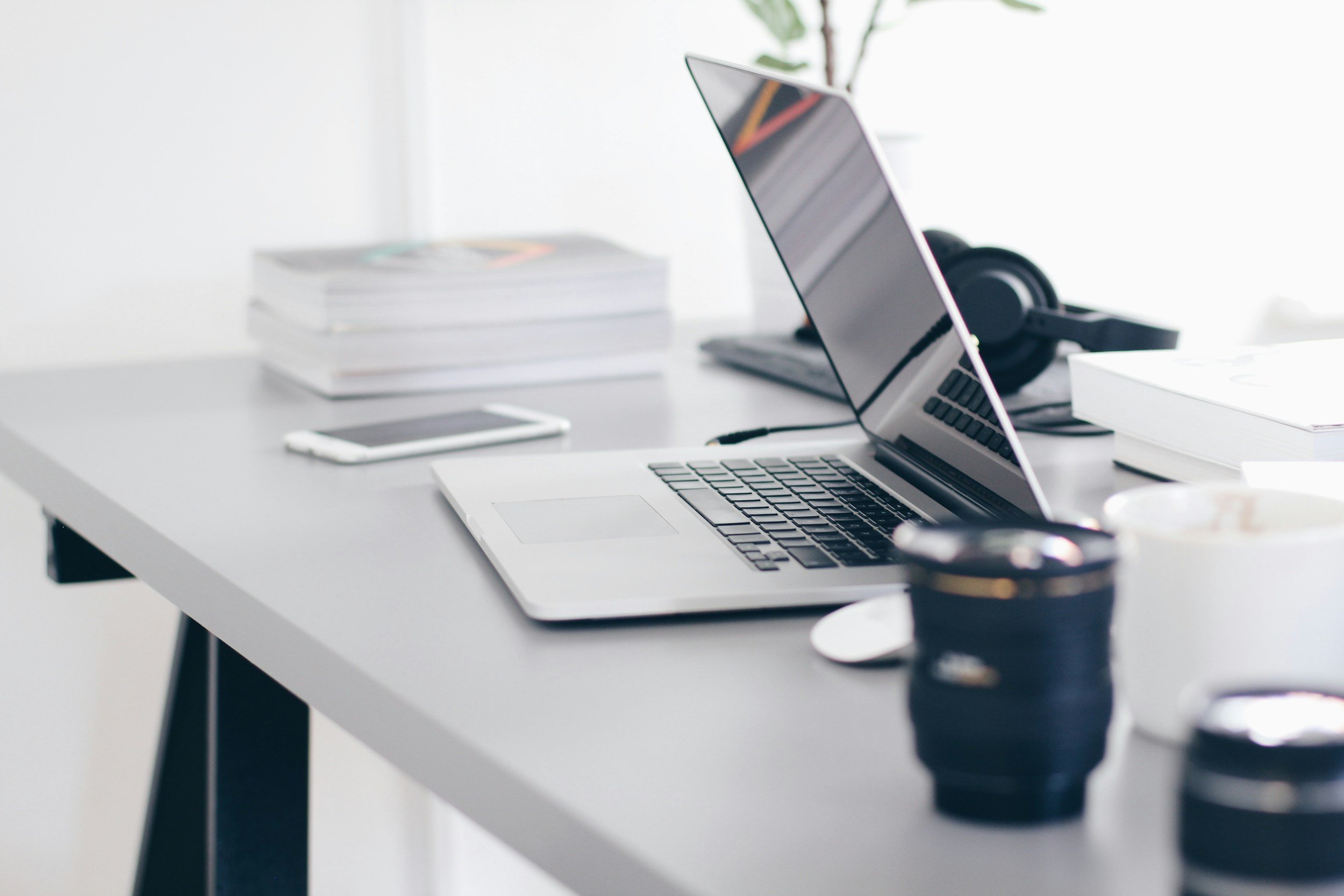 Desk with an open laptop, smartphone, camera lens, headphones, stacks of books, and a cup, in a bright workspace.