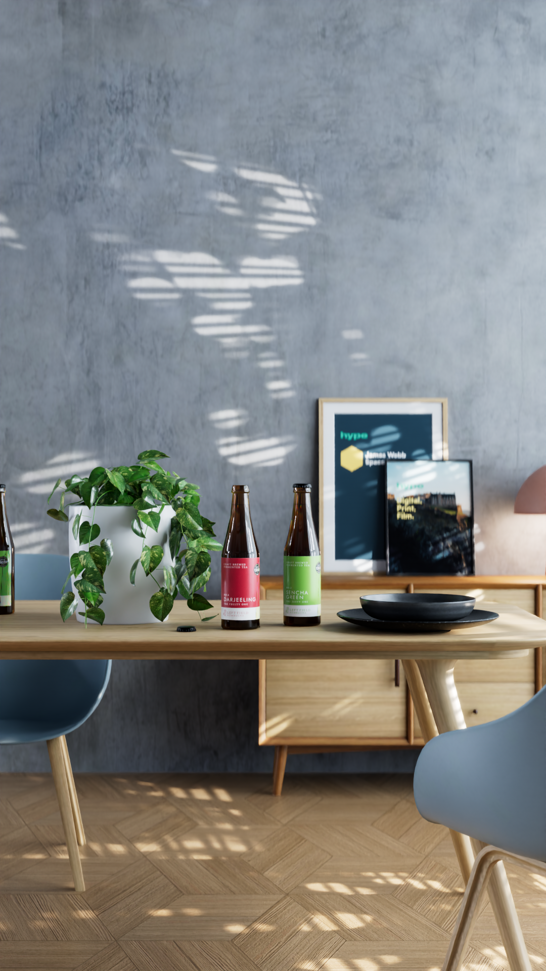 A wooden dining table with potted plant, three bottles of tea, black plates, and framed artwork against a gray textured wall with sunlight patterns.