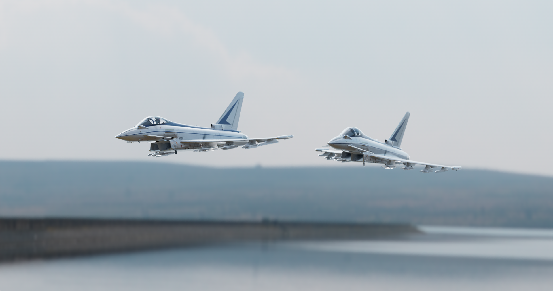 Two fighter jets flying in the sky over a body of water with a shoreline visible in the background.