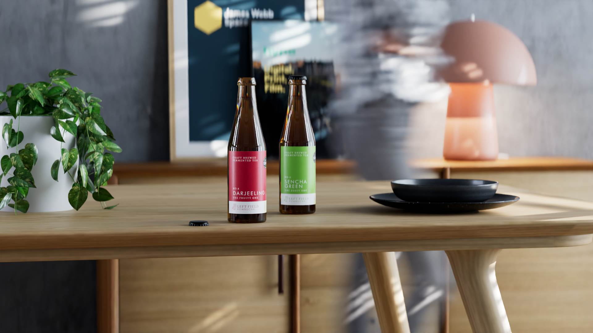 Two bottles of craft brewed fermented tea on a wooden table with a black plate and a potted plant nearby. A blurred background includes a framed picture and a pink lamp.