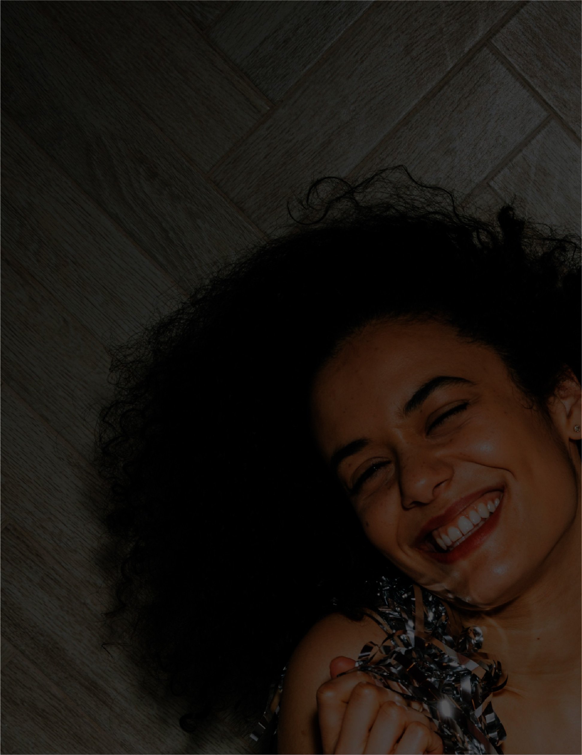 Close-up of a woman with curly hair laughing and holding metallic confetti, with a wooden floor background.