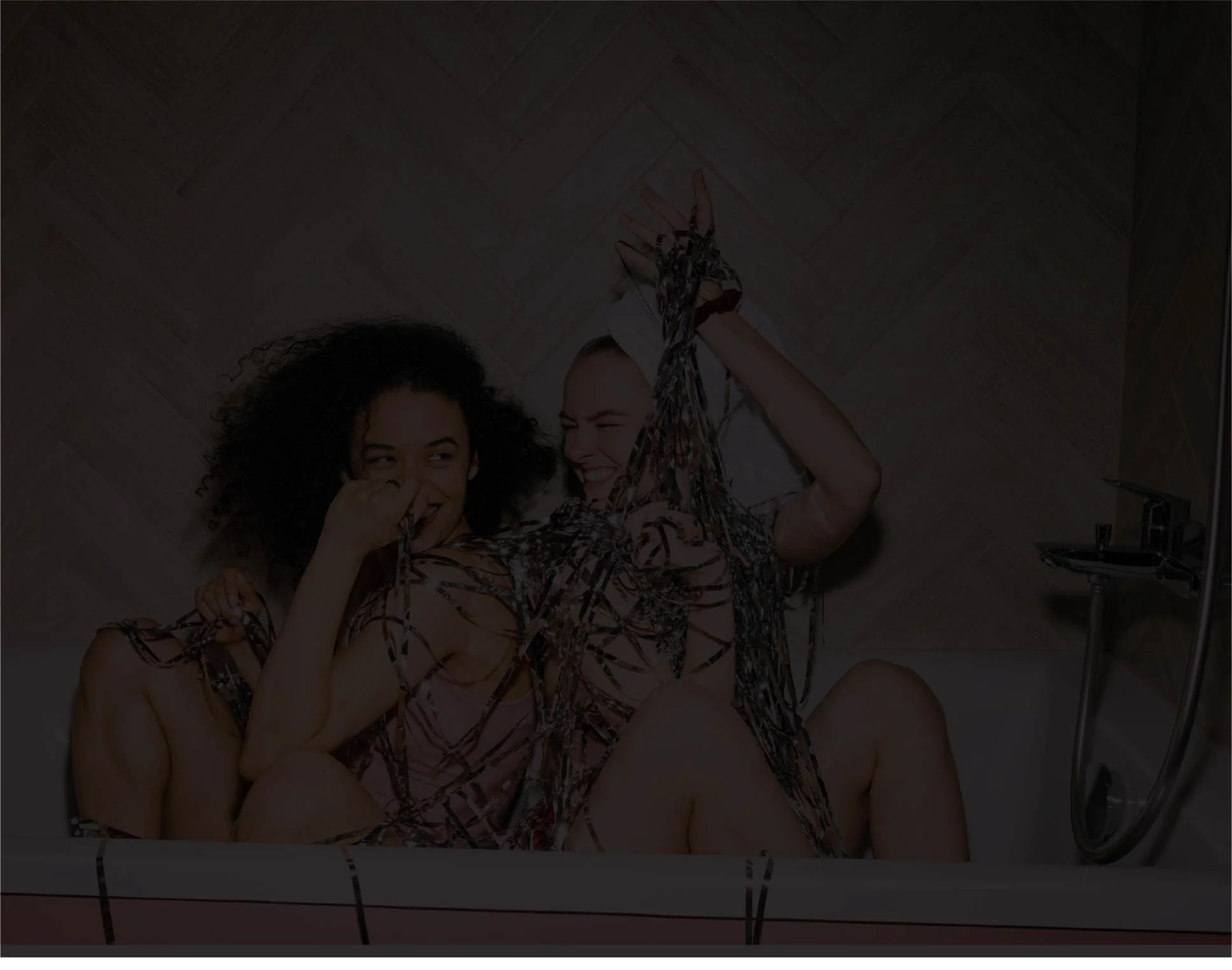 Two women are lying on a bathroom floor covered with metallic streamers. They are smiling and looking at each other. One woman has curly dark hair and the other woman has lighter hair, both appearing to be having fun.