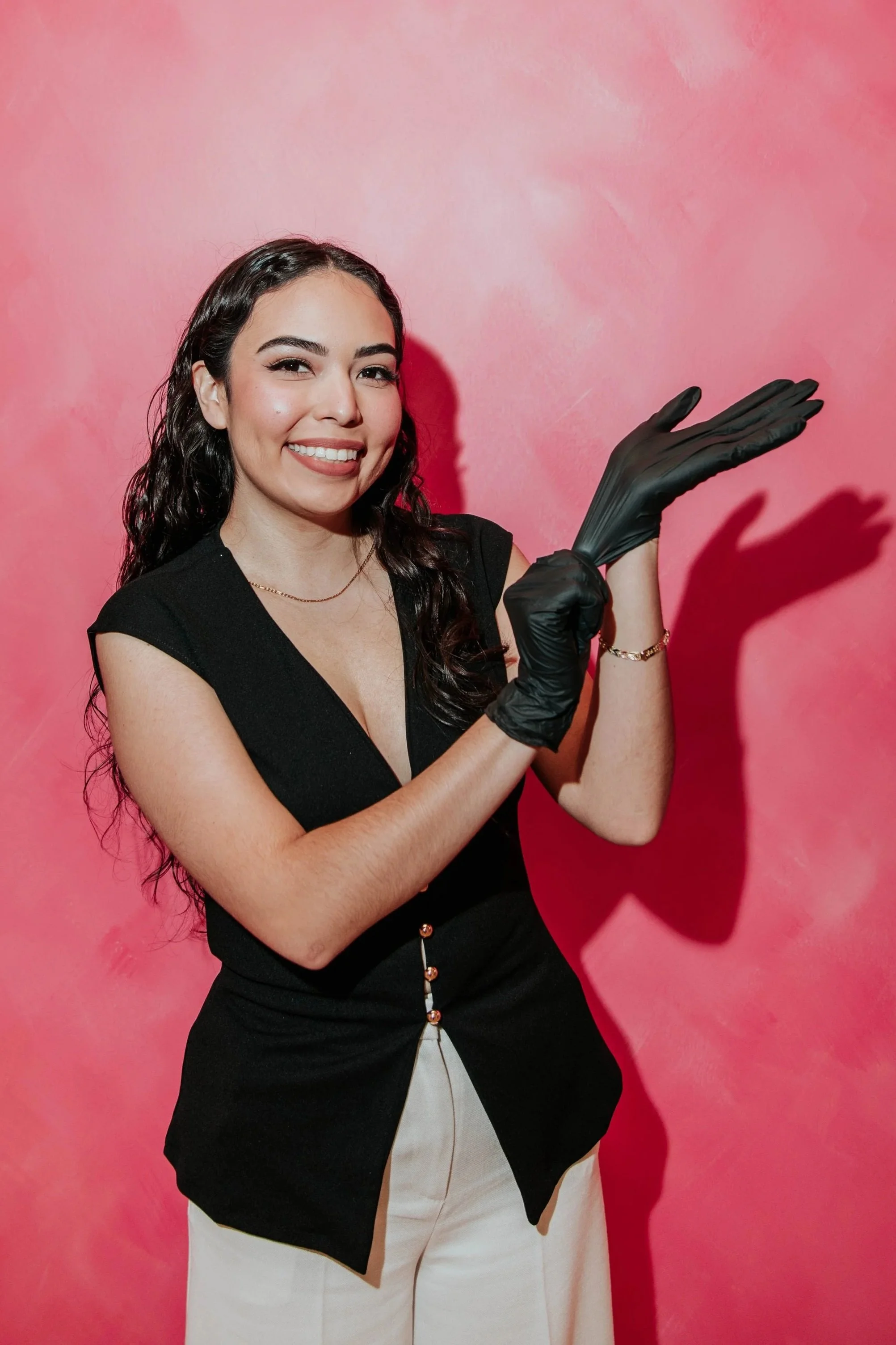 A smiling woman with dark, wavy hair, wearing a black sleeveless top, beige pants, and black gloves, standing against a pink background.