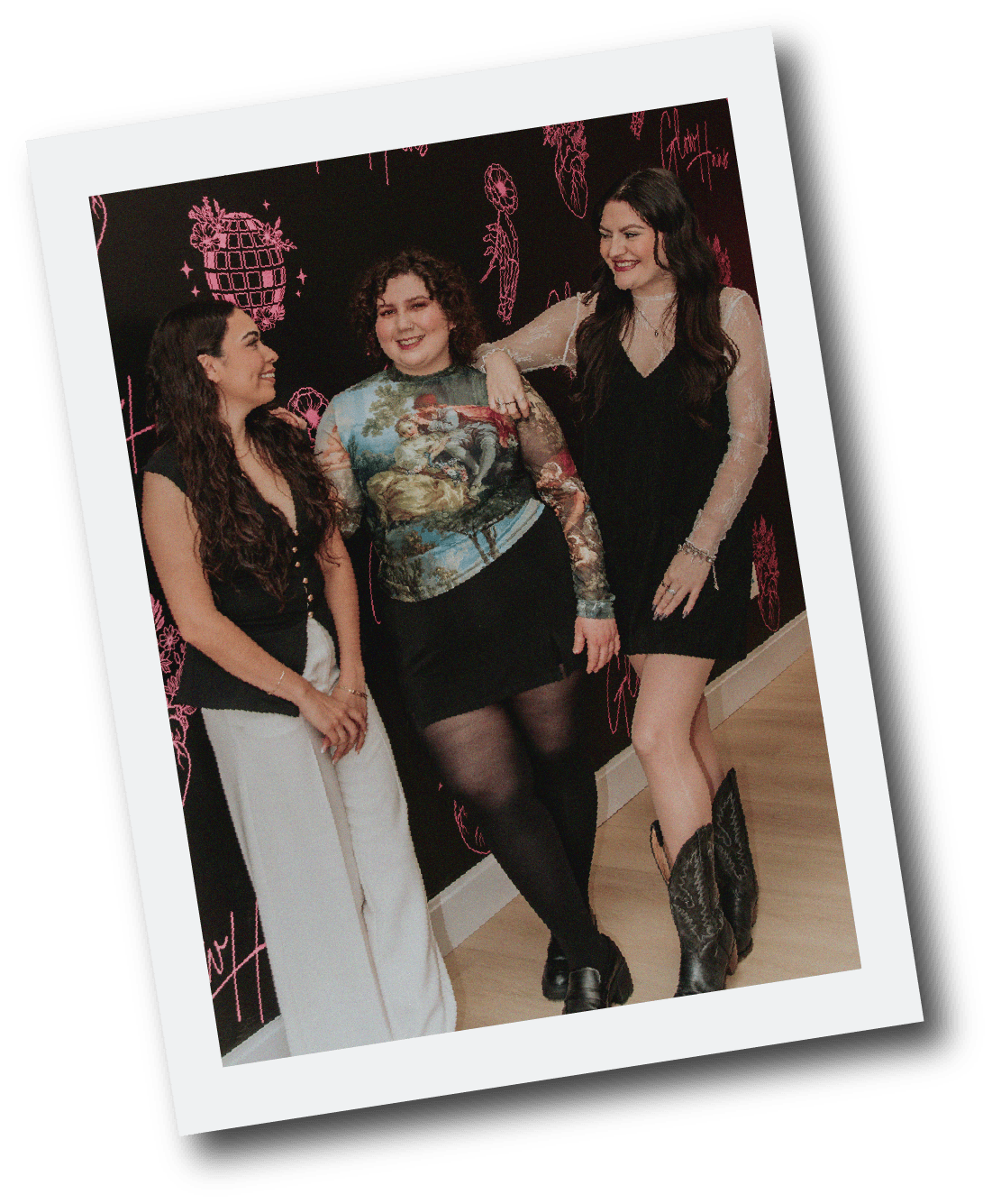 Three women smiling and conversing at an event, standing against a black wall with pink graphic designs, including disco balls and skeleton hands, with a white frame tilted at an angle.