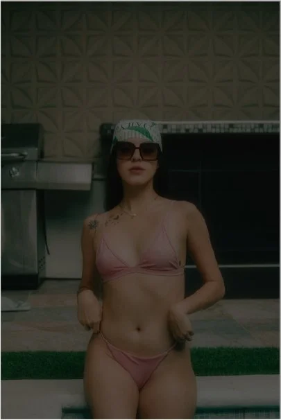 A woman in a pink bikini, wearing sunglasses and a white cap, standing indoors in front of a patterned wall and a piano.