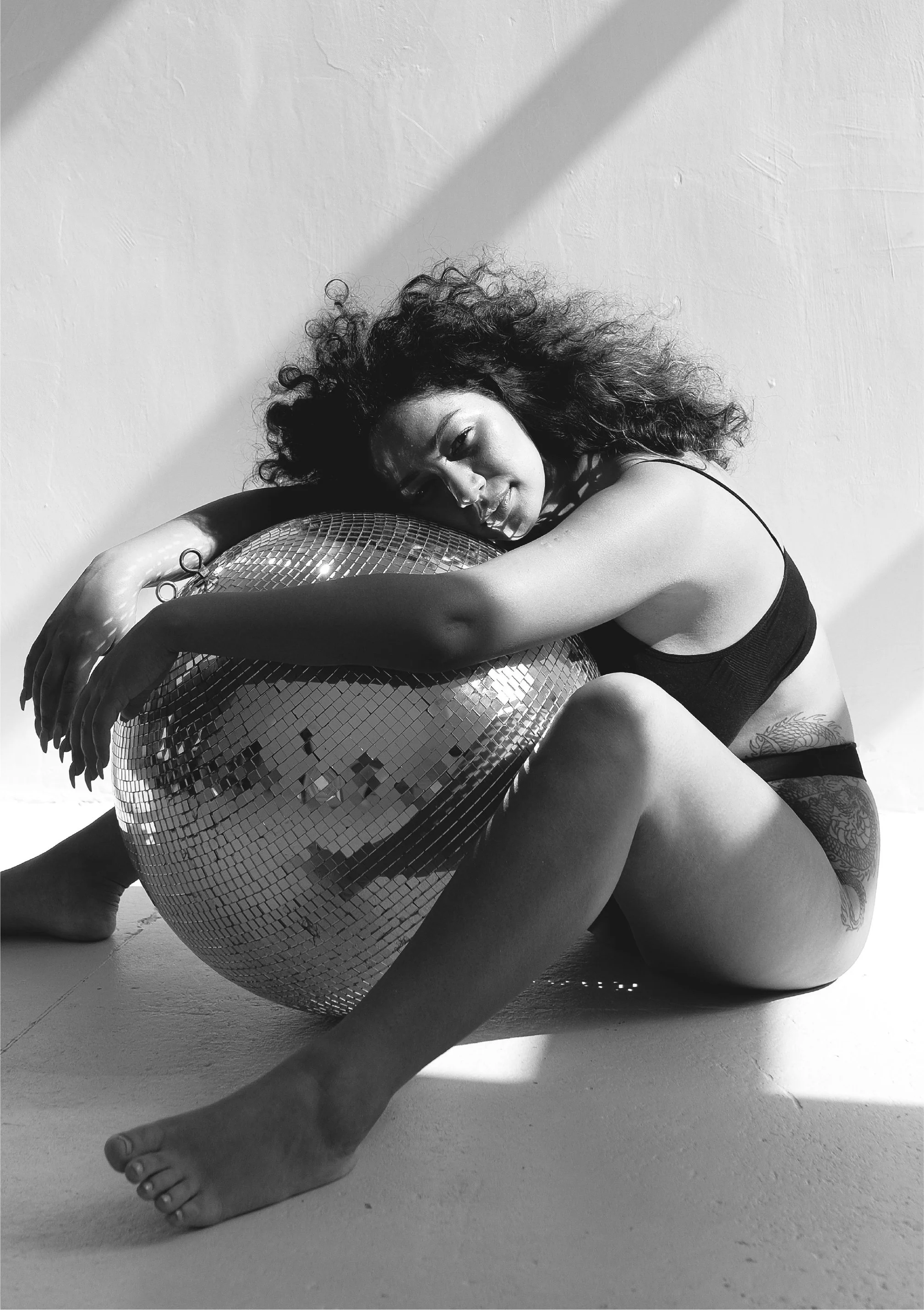 Black and white photo of a woman with curly hair, resting her face and arms on a large disco ball, on the floor, against a plain wall.
