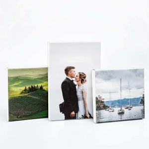 Three framed pictures leaning against a white background. The left picture shows a green landscape with rolling hills, the middle picture features a bride and groom in wedding attire, and the right picture depicts boats docked at a marina on a cloudy day.