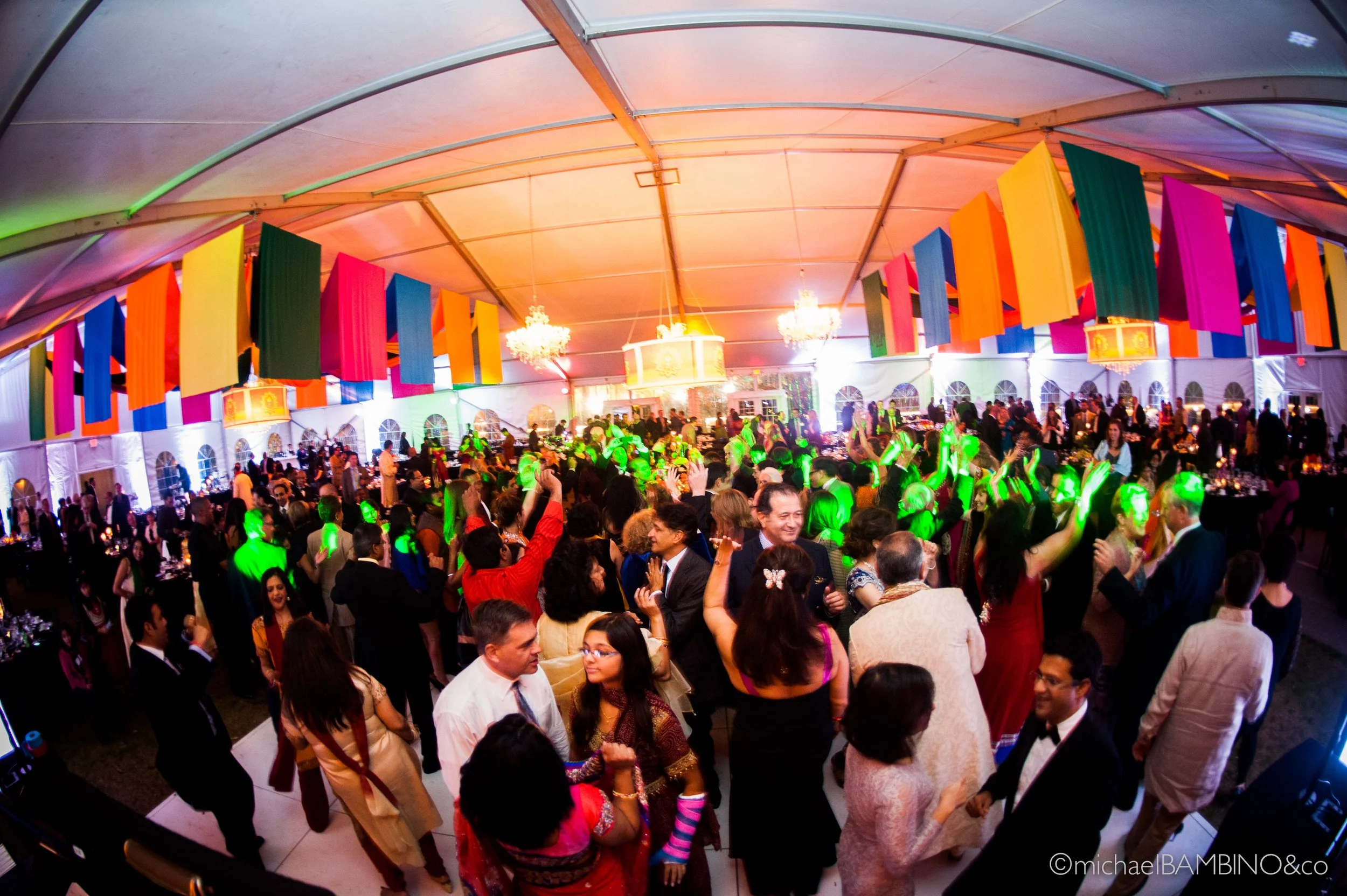 A lively indoor celebration with colorful banners hanging from the ceiling, chandeliers, and a crowd of people dancing and socializing under vibrant lighting.