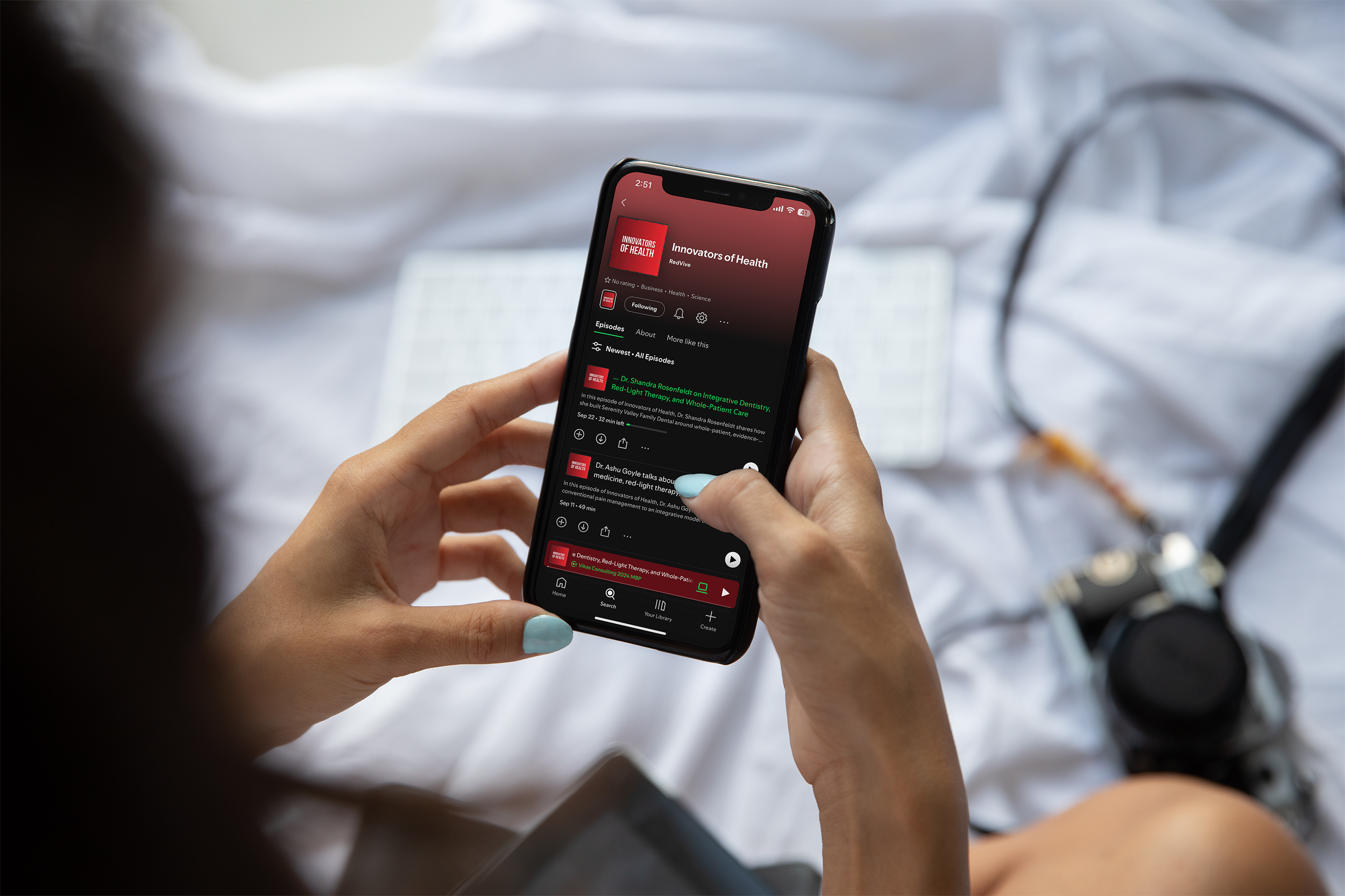 Person holding a smartphone with the 'Innovators of Health' podcast displayed on the screen, sitting on a bed with a camera and a notepad nearby.