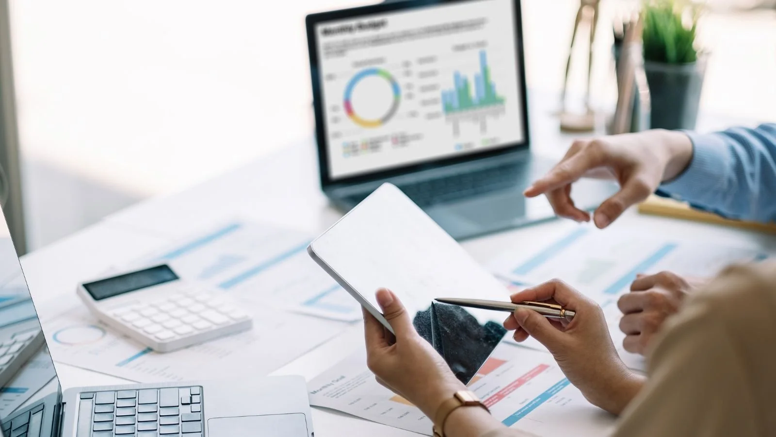 Business meeting with financial charts on laptop and papers on table, person holding smartphone and pointing at a document.
