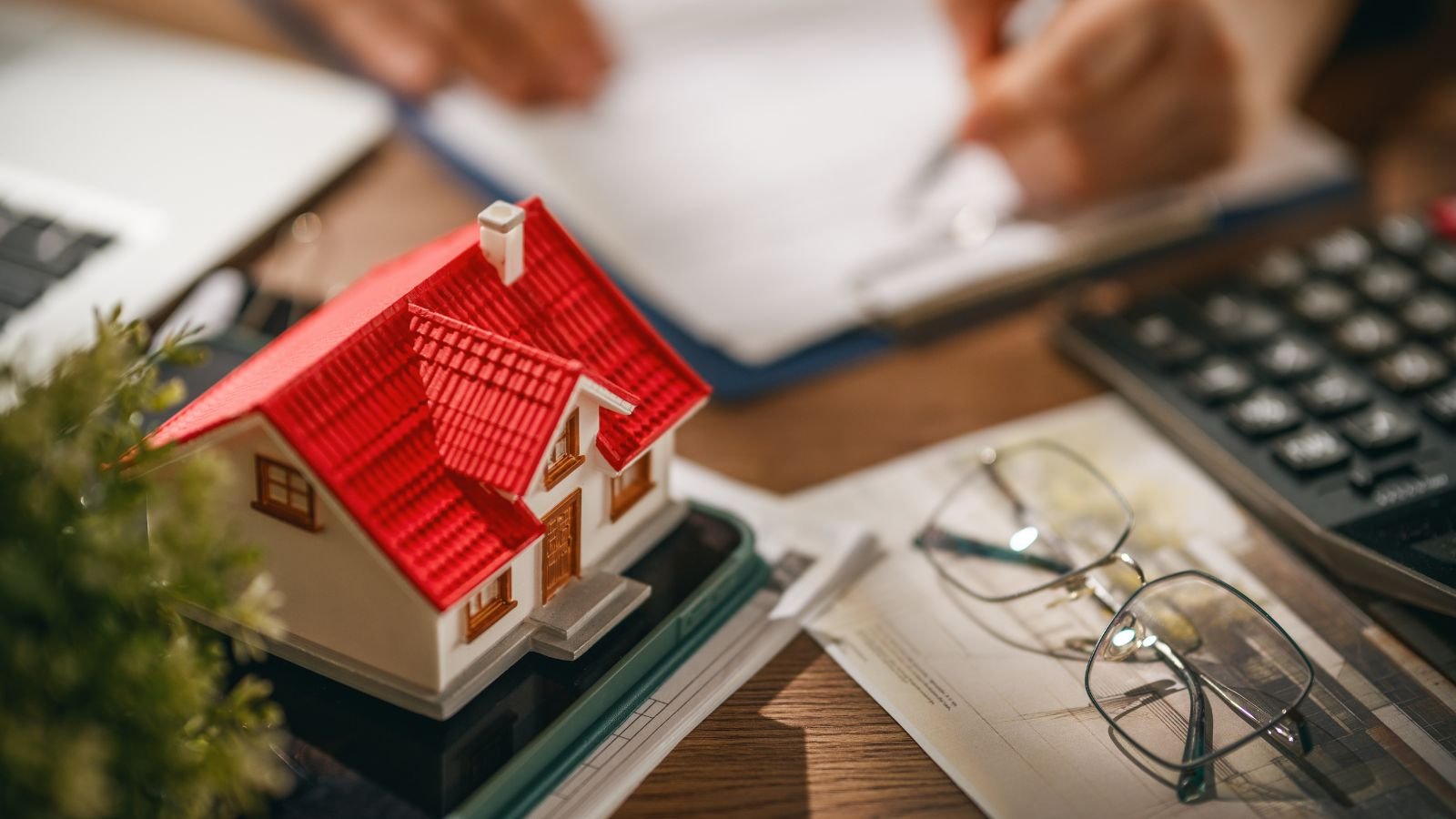 A miniature model house with a red roof on a desk, surrounded by papers, eyeglasses, a calculator, and a person writing in a notebook in the background.