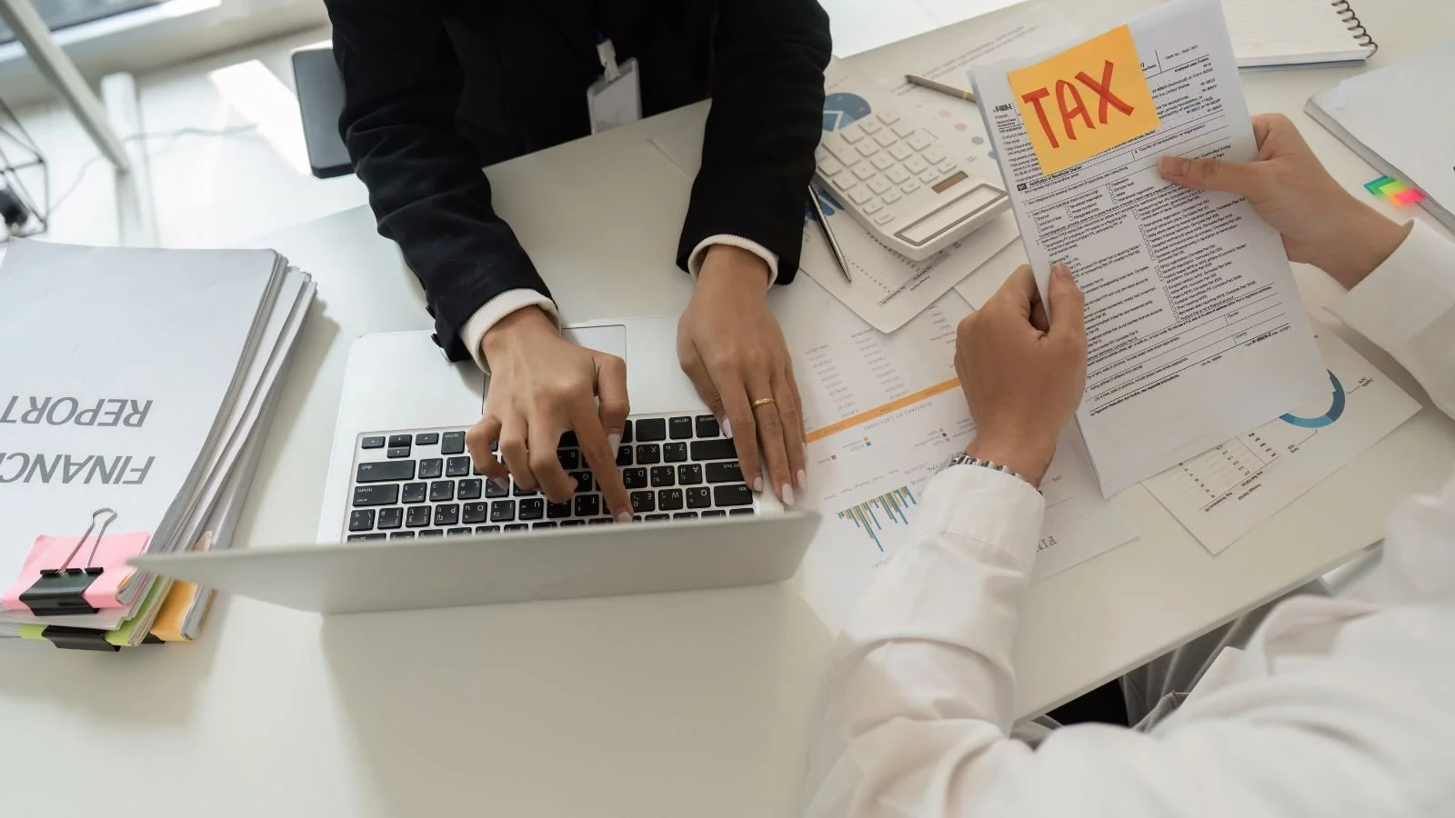 Two people reviewing tax documents at a desk, one pointing at the laptop keyboard, the other holding a paper labeled 'TAX' with sticky notes.