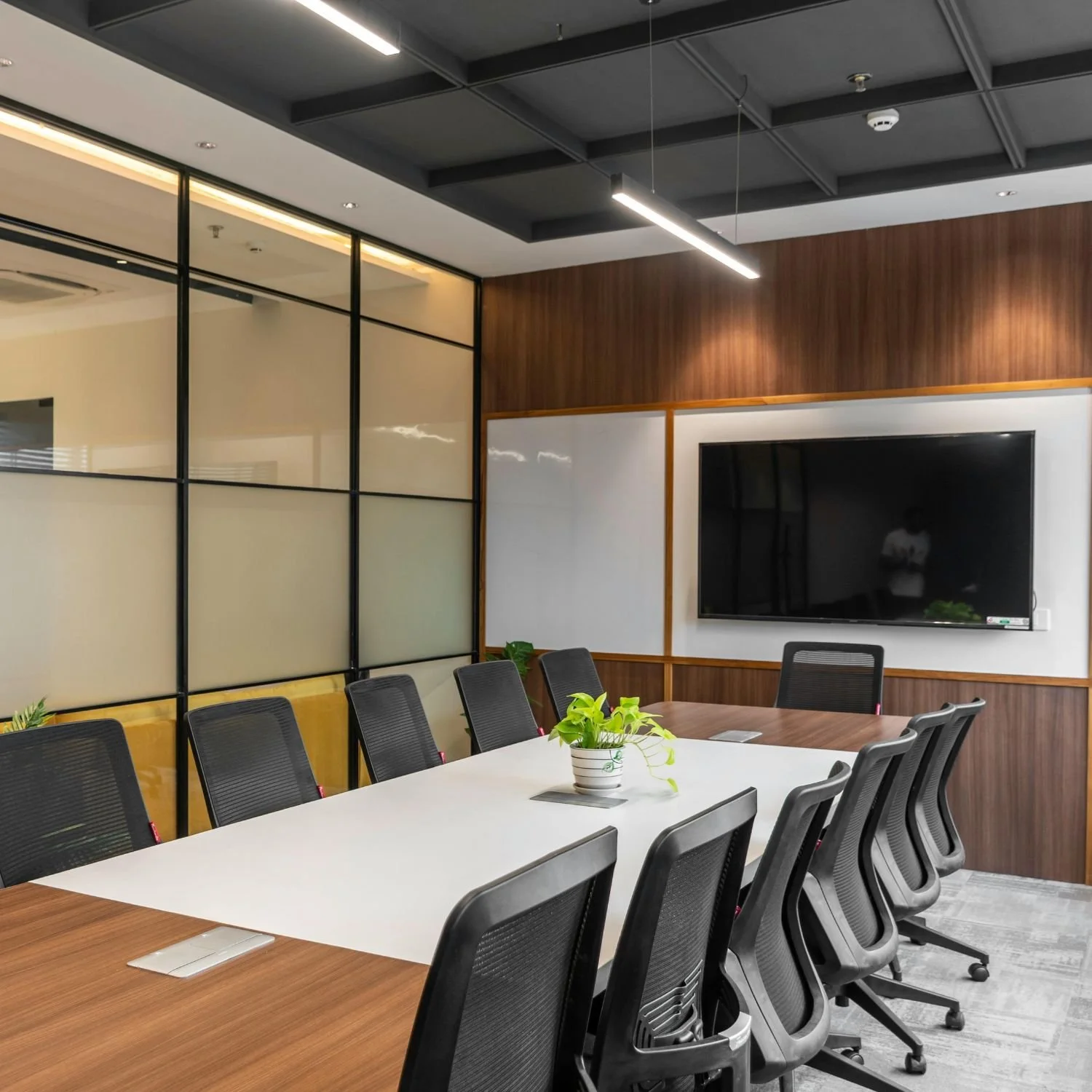 Modern conference room with a long white table, black chairs, a potted plant, a large flat screen TV, wood paneling, frosted glass partition, and ceiling lighting.